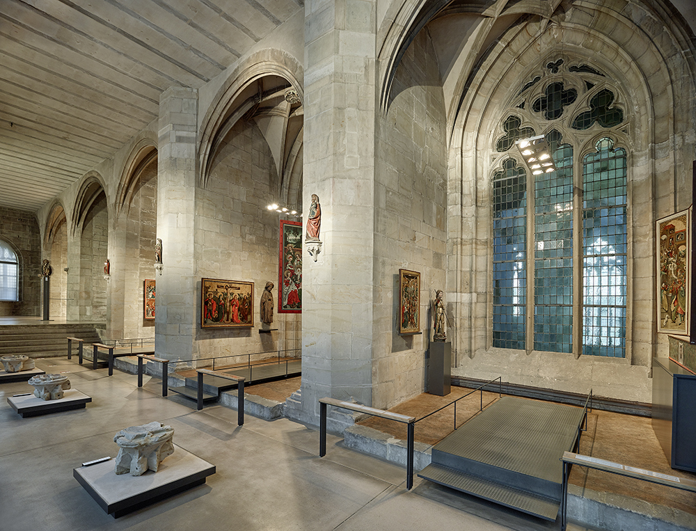 Kapitelsaal mit Altarbildern. Ein langgezogener Raum mit Nischen, in denen mittelalterliche und neuzeitliche Altarbilder hängen. Eines der großen gotischen Fenster ist erkennbar und gibt den Blick auf das Mittelschiff des Domes frei. Vor den trennenden Wänden hängen bunte Skulpturen von Heiligen. Davor stehen architektonische Baukörper des Domes, wie Schlusssteine und Kapitelle.