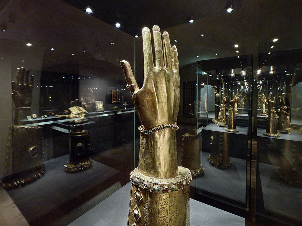 Die Schatzkammer. Im Vordergrund steht ein goldenes Reliquiar in Form einer erhobenen Hand. Am Handgelenk befindet sich ein edelsteinbesetztes Armband und am Daumen ein Ring. Durch die Glasvitrine sind weitere goldene Armreliquiare, Bücher und andere Schätze im Hintergrund erkennbar.