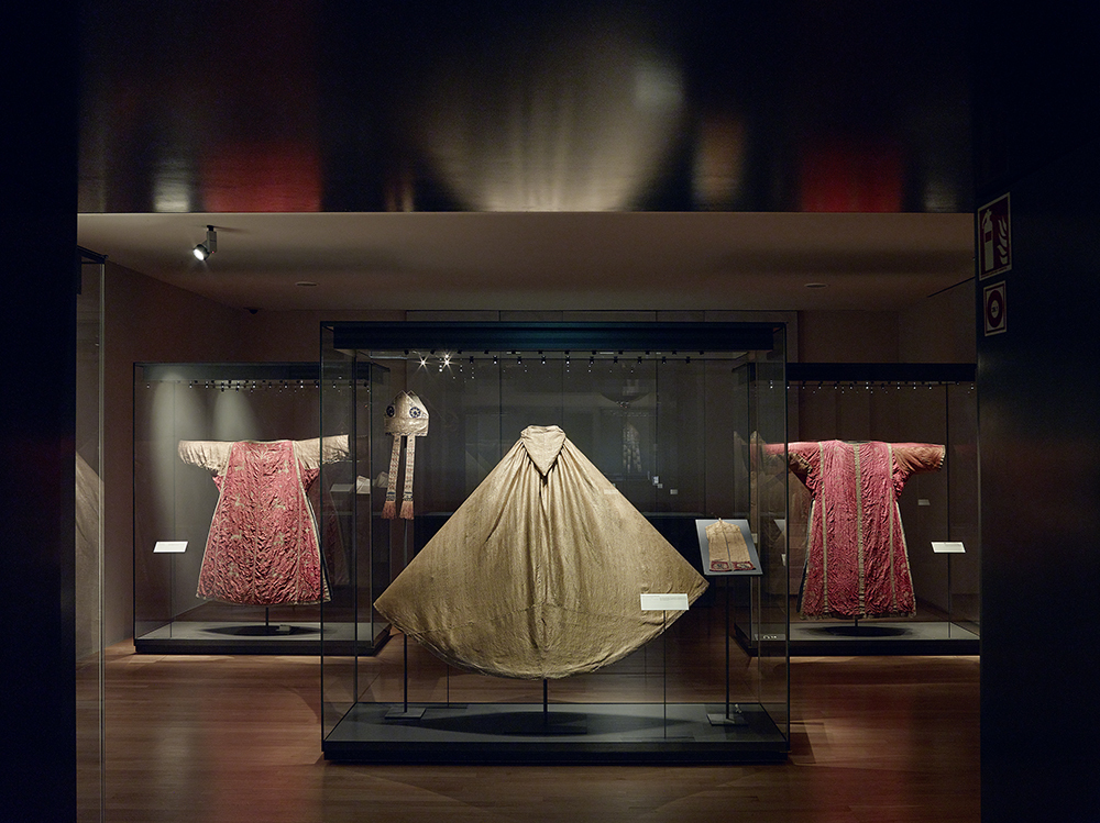 Im Gewändersaal wird ein goldener Chormantel aus Seide präsentiert. Daneben liegt eine goldene Stola und eine bestickte Mitra des Bischofs. Die Vitrine wird flankiert von zwei roten Dalmatiken aus Seide und Goldstickerei.