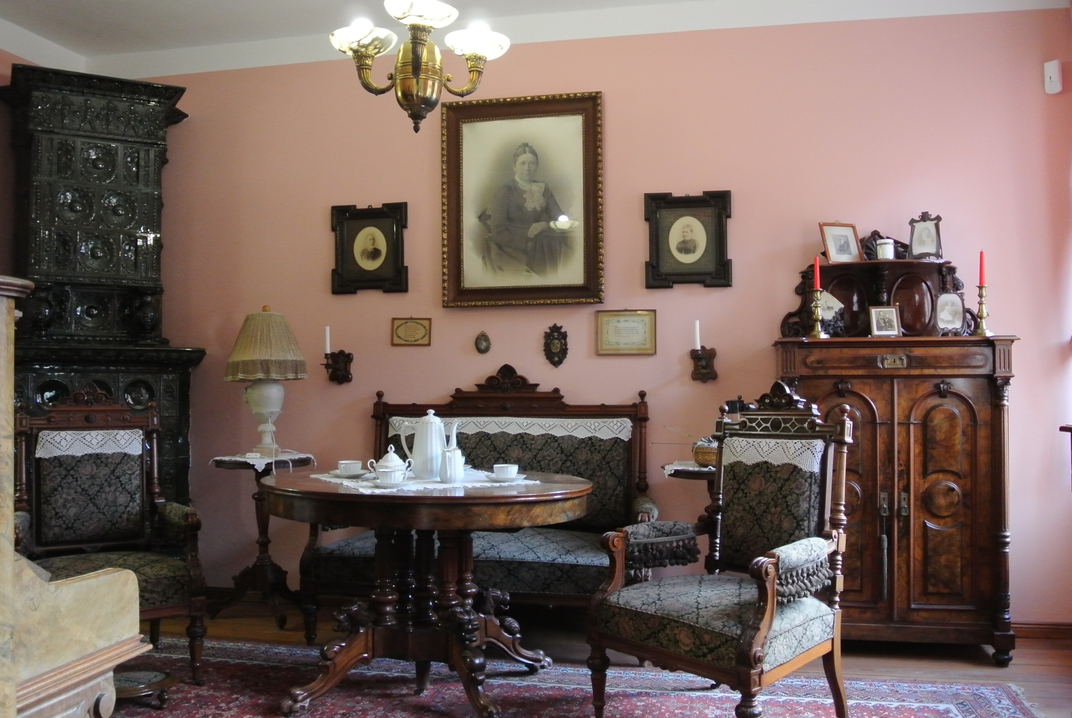 In der Stube der Familie stehen dunkle Holzmöbel um einen runden Tisch mit weißem Kaffeegedeck. In der Ecke steht ein dunkler Kachelofen. An der hellrosa Wand hängen einige eingerahmte Porträts der Familie.