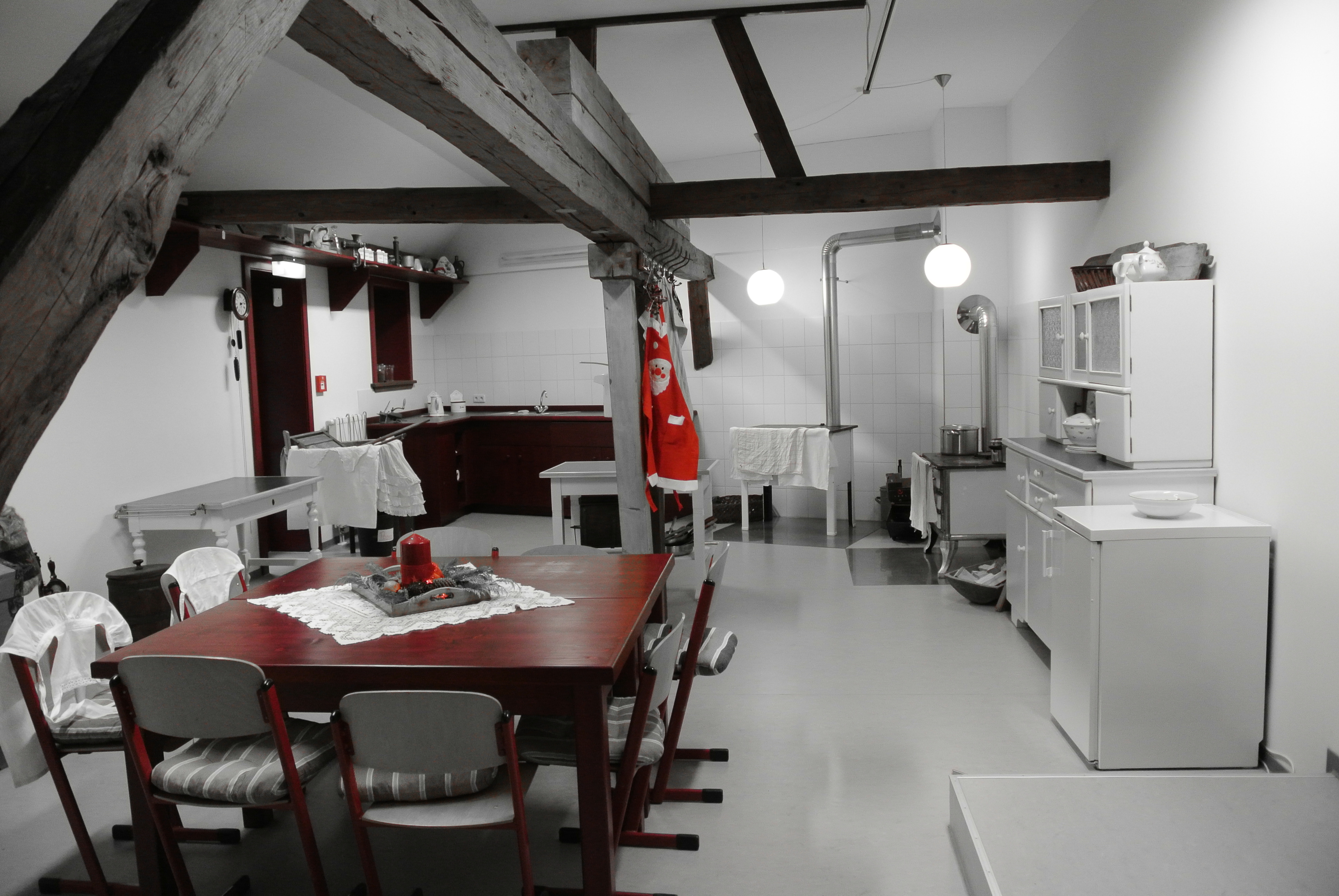 Im Vordergrund steht ein Holztisch. Eine Küche mit Kohleöfen befindet sich im Hintergrund. Das Foto ist schwarz-weiß, bis auf einige rote und dunkelbraune Elemente.