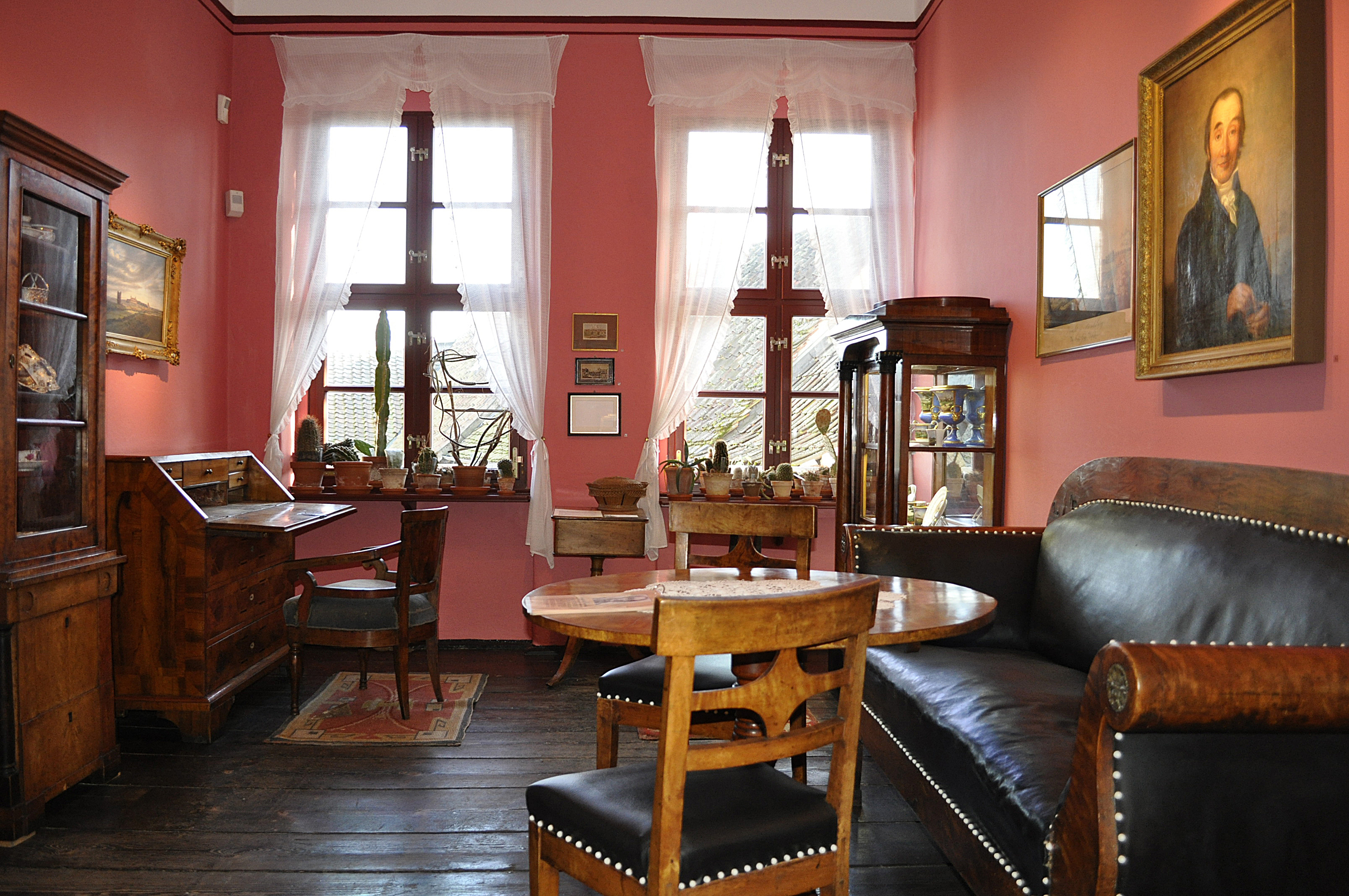 Dauerausstellung Biedermeierzeit und J. G. Nathusius. Ein hellrotes Zimmer mit einem dunklen Holzfußboden, das mit Interieur aus der Biedermeierzeit ausgestattet ist. Ein großes mit schwarzem Leder bezogenes Sofas mit passender Bestuhlung stehen um einen runden Holztisch. in dunkler Schreibtisch und eine Vitrine mit Kunsthandwerk stehen vor zwei großen Fenstern im Hintergrund.