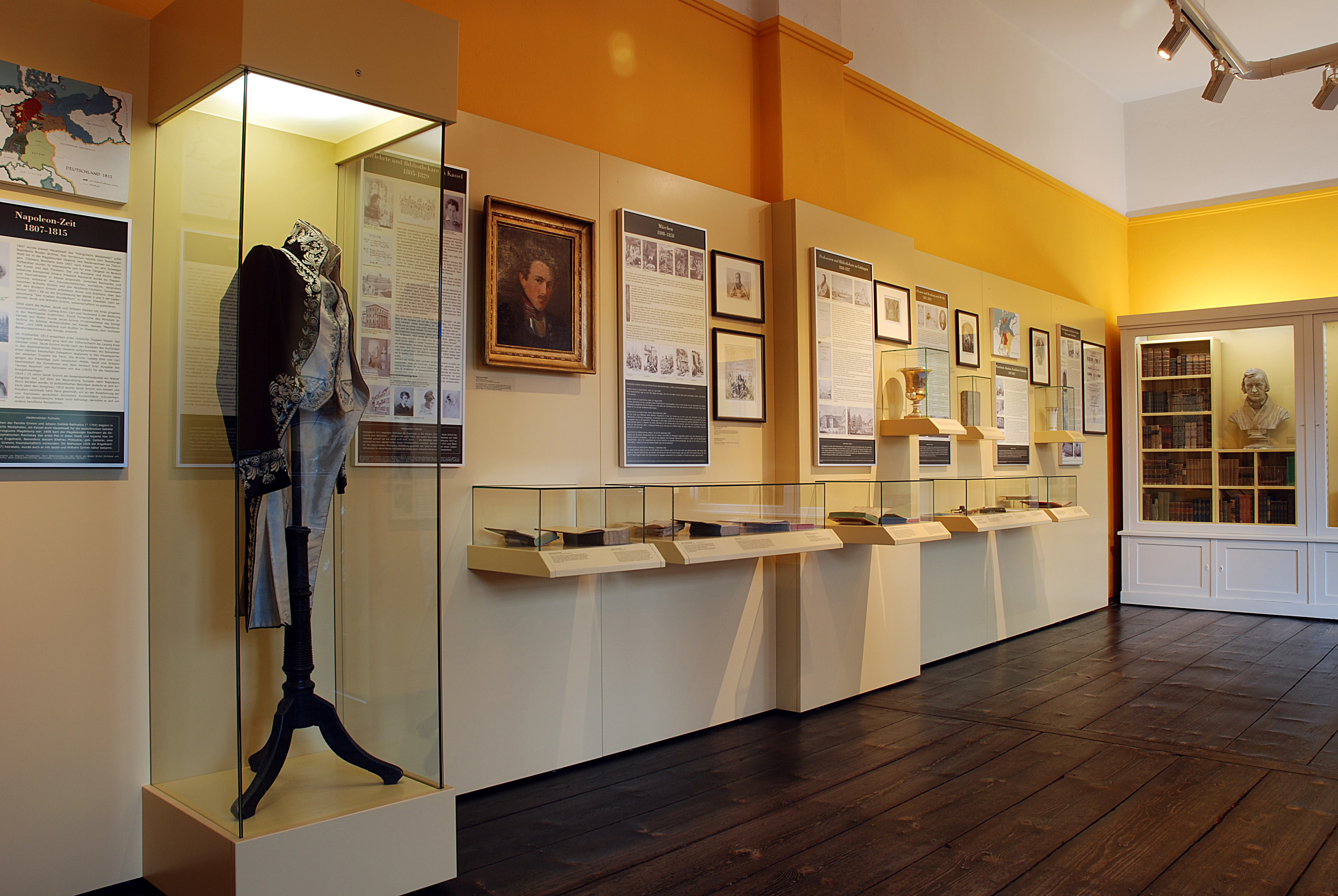 In der Dauerausstellung "Brüder Grimm" fällt der Blick direkt auf eine hohe Vitrine, in der ein blau-weißer Frack ausgestellt wird. Des Weiteren hängen längliche Vitrinen mit Büchern sowie Texttafeln und Bilder an der Wand. Ein weißes Bücherreal, das auch eine helle Büste beherbergt, steht am Ende des Raumes.