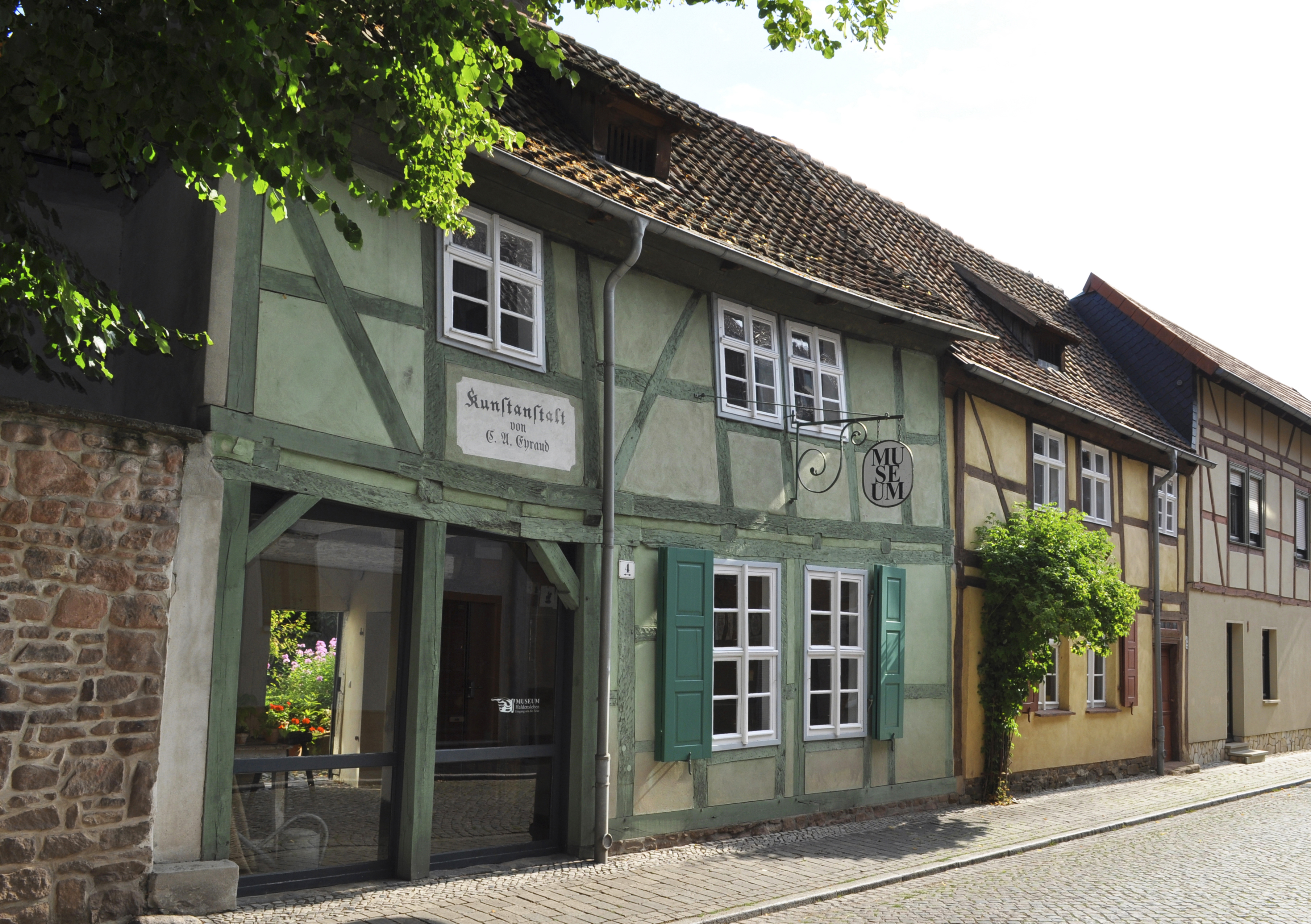 Fachwerkhäuser Lange Straße 2 und 4. Neben einem grünen stehen zwei gelbe Fachwerkhäuser. Über einem großen Schaufenster des grünen Hauses wurde ein Schild mit der Aufschrift "Kunstanstalt" aufgemalt. Ein weiteres Schild hängt über dem Gehweg und verrät, dass es sich hier um ein Museum handelt. Zwischen dem grünen und gelben Haus befindet sich ein hoher Rosenbusch.