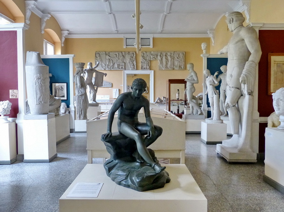 In der Ausstellung des Archäologischen Museums sind viele Gips-Abgüsse antiker Statuen und anderer Objekte. Eine Bronze-Statue sitzt in der Mitte des Raumes auf einem Stein und streckt das Bein aus. Die Abgüsse umgeben sie von beiden Seiten.