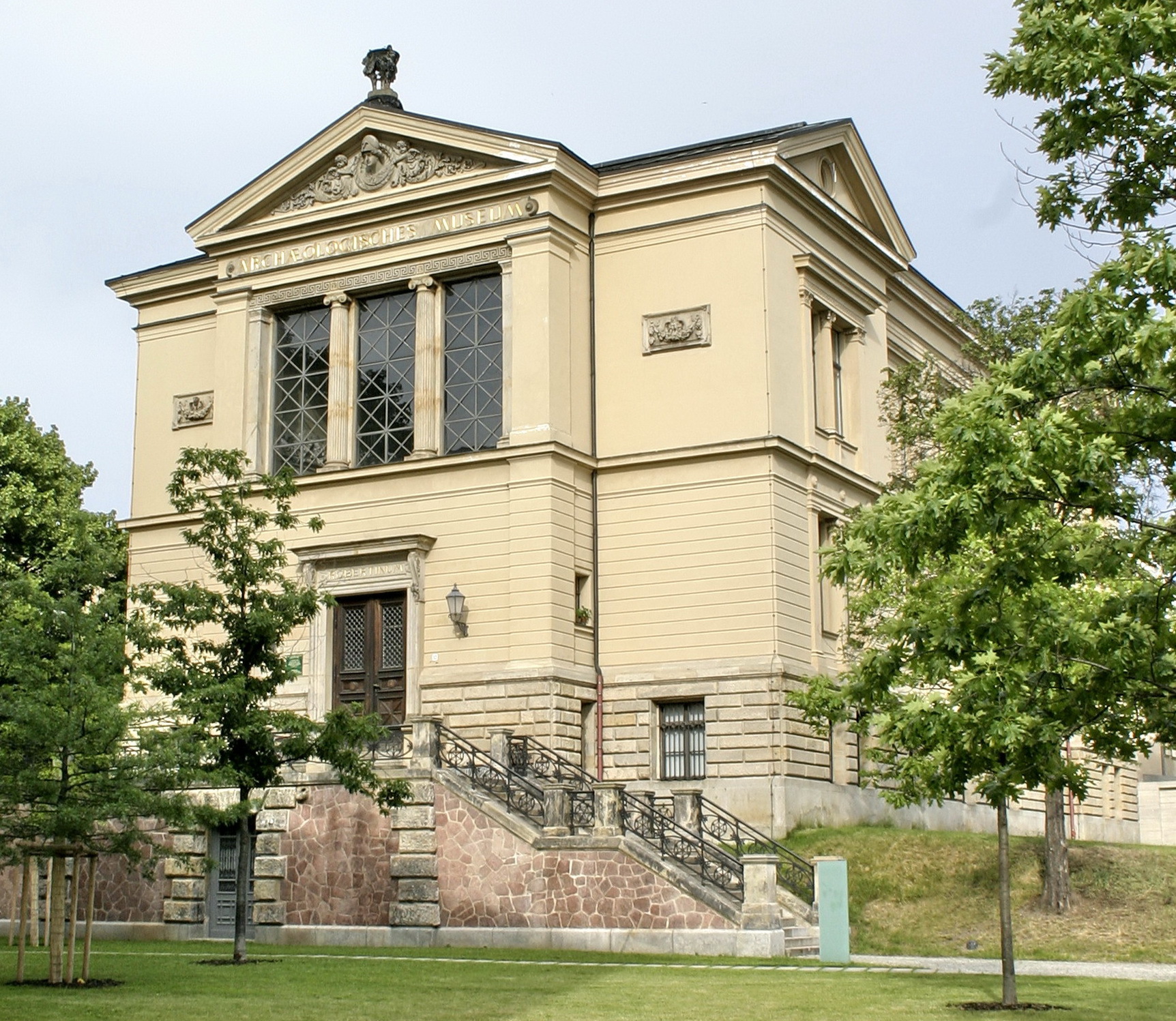 Das Robertinum von Außen. Vor dem Gebäude stehen kleine Bäume auf einer Wiese. Eine Steintreppe führt hoch zum Eingang. Die Fassade ist hellgelb und gliedert sich in einen Mittelrisalit mit drei großen Fenstern und einem Tympanon. Die Seiten werden flankiert von zwei Steintafeln mit dekorativen Girlanden.