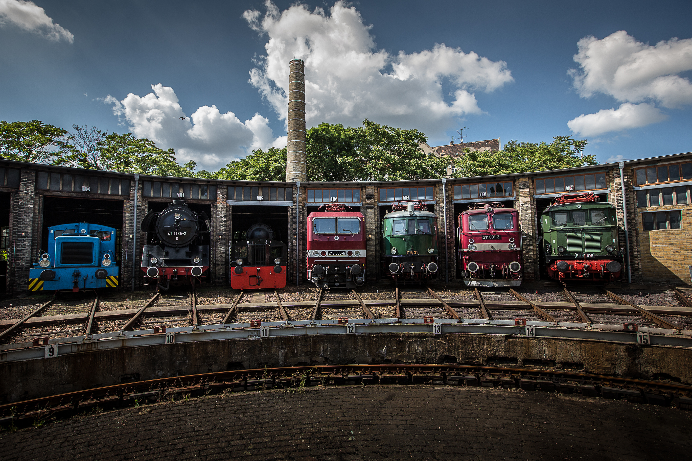 Sieben bunte Bahnen stehen aufgereiht in ihren Schuppen und schauen mit der Vorderseite heraus. Ein Gleisbett befindet sich vor ihnen.