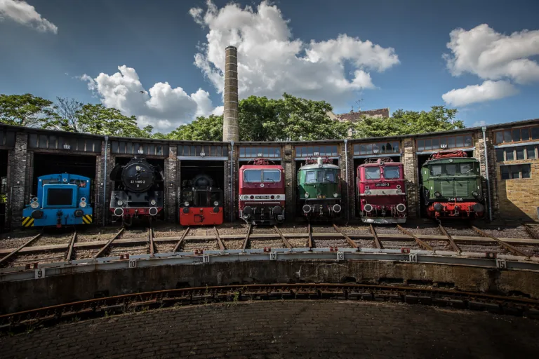 Sieben bunte Bahnen stehen aufgereiht in ihren Schuppen und schauen mit der Vorderseite heraus. Ein Gleisbett befindet sich vor ihnen.