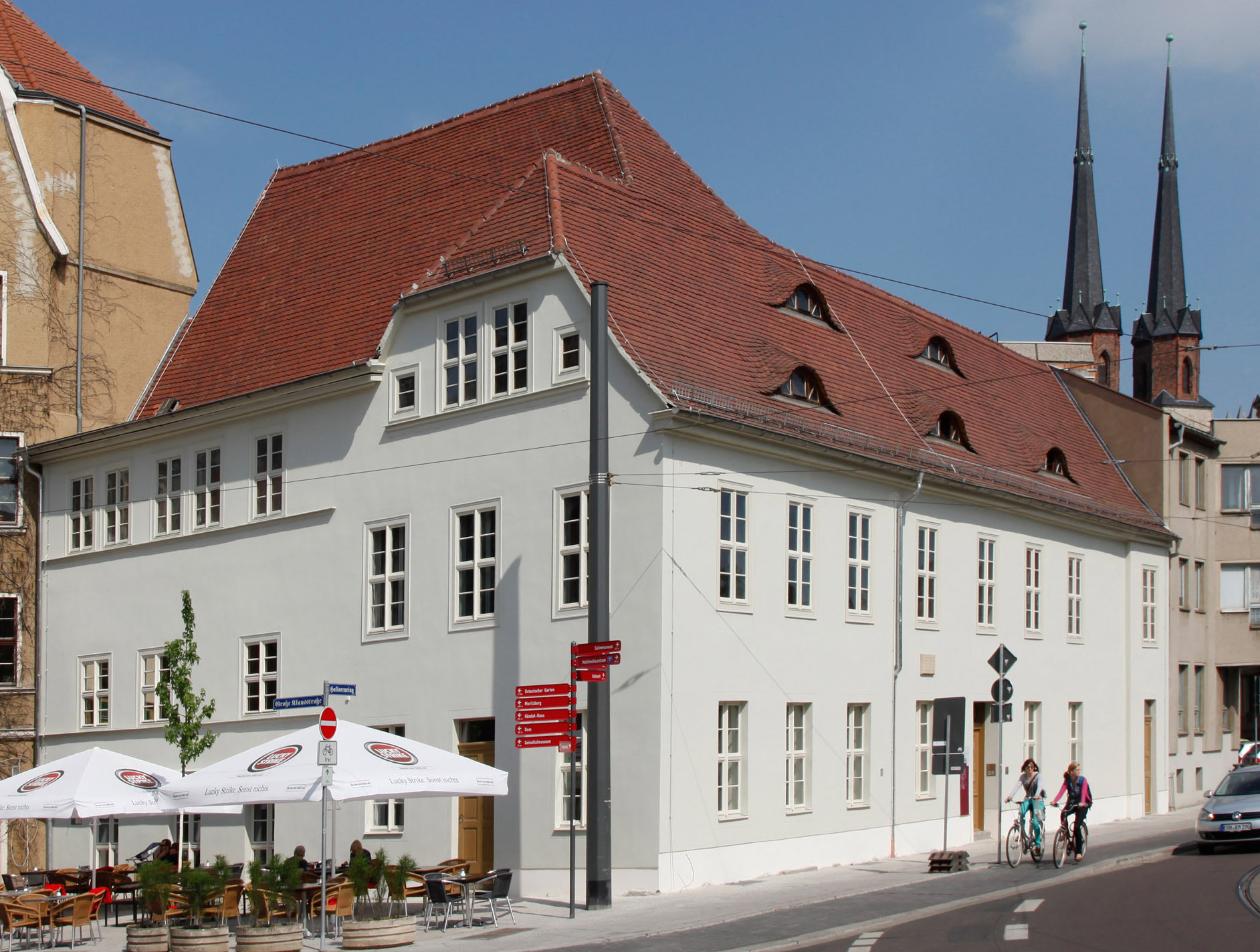 Die hellgrüne Fassade des Wilhelm-Friedemann Bach-Hauses. Davor stehen Tische und Stühle mit weißen aufgespannten Schirmen. Im Hintergrund sind zwei Türme der Marktkirche Halle zu erkennen.