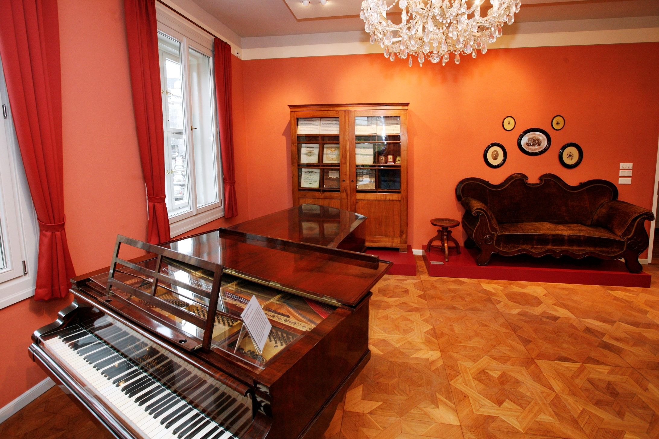 Im Vordergrund des Robert Franz Salons steht ein geöffnetes Klavier. Aufgrund einer Glasplatte auf der Oberseite sind die Saiten des Instrumentes erkennbar. Vor den rot gestrichenen Wänden steht ein dunkles Sofa und ein Holzschrank, in dem Notenblätter zu erkennen sind.