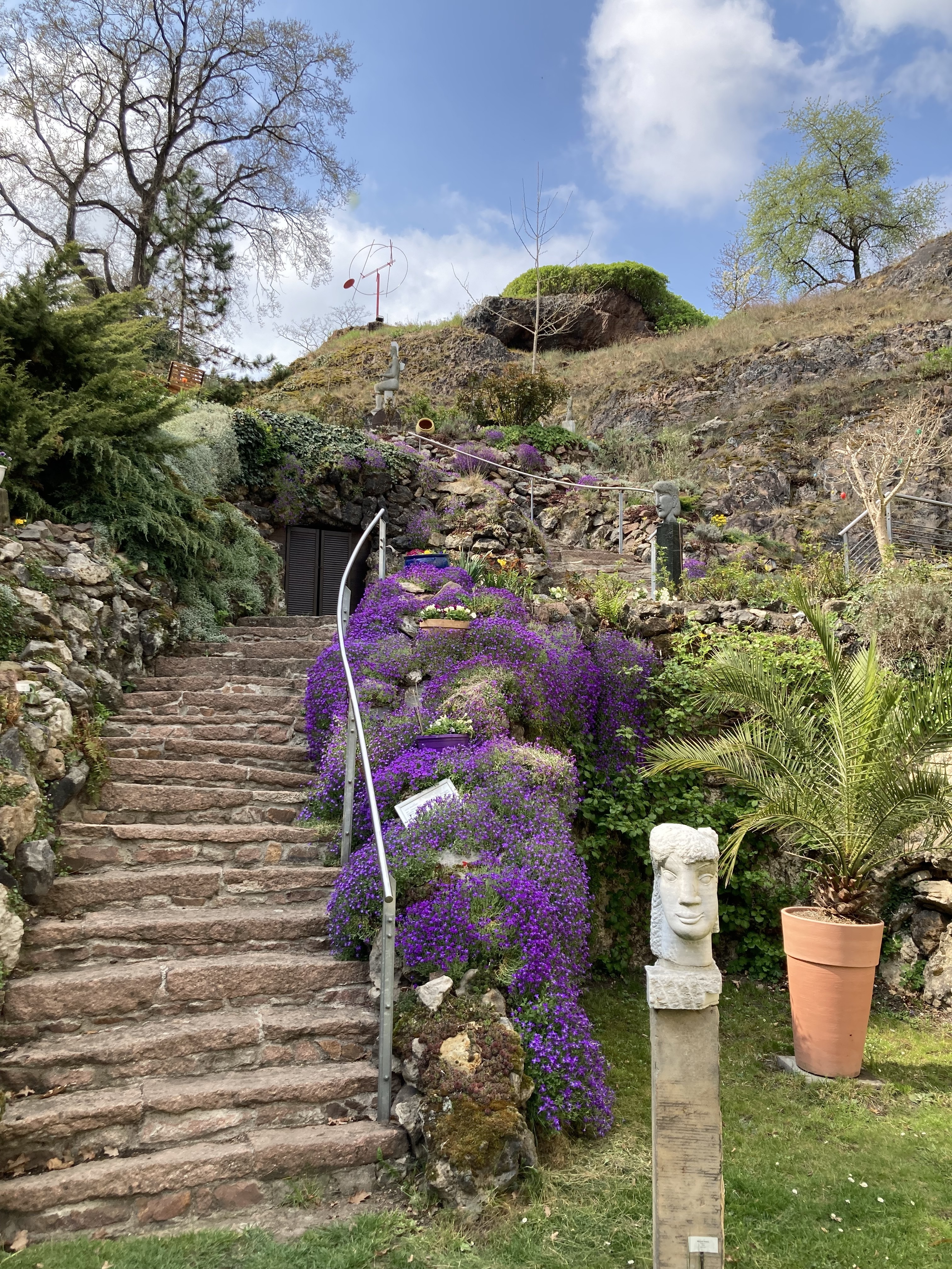 Eine Treppe aus Stein führt in den Felsengarten herauf. An der Seite wachsen lila und gelbe Blume. Vor der Treffe stehen eine  Palme und eine Büste.
