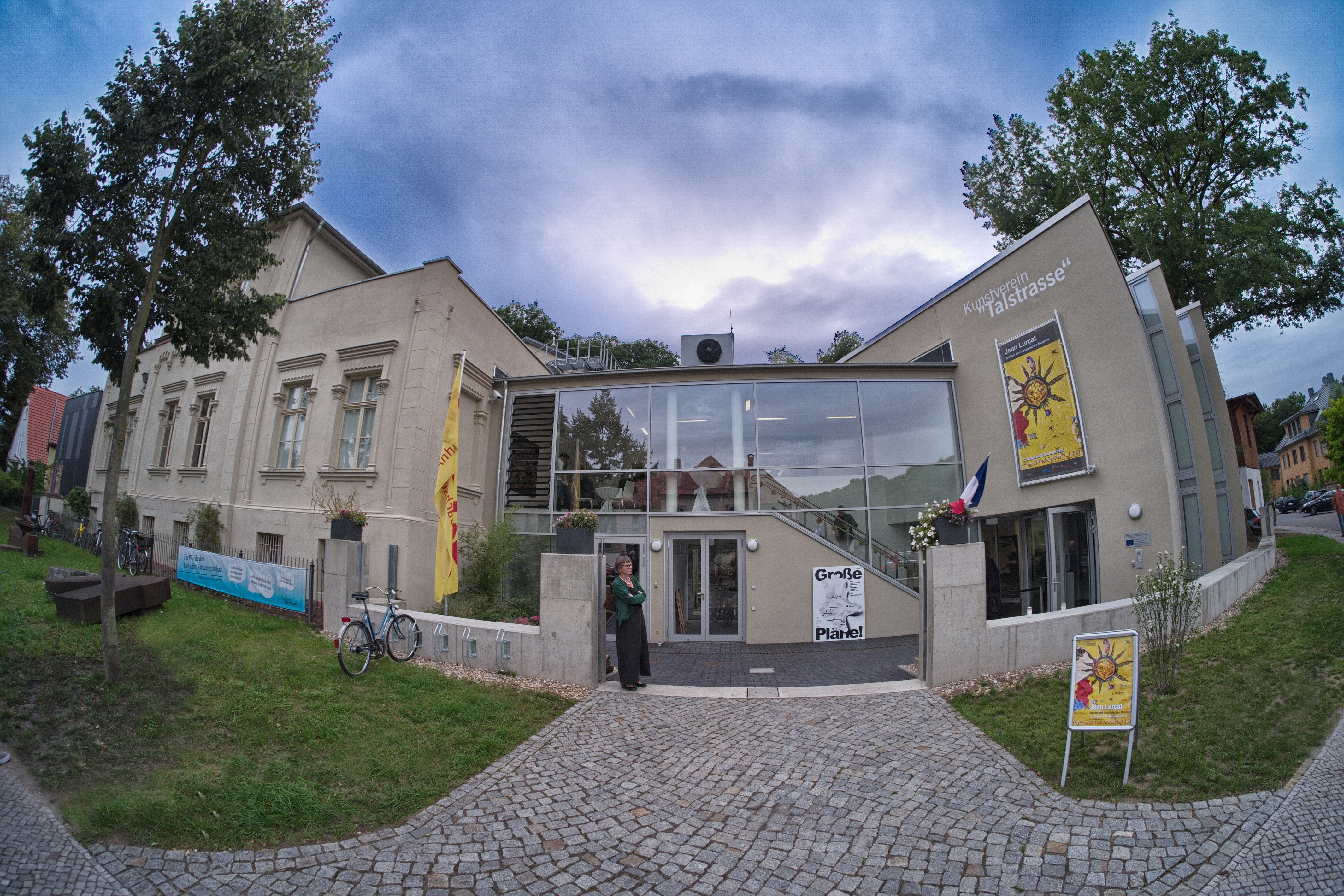 Die Kunsthalle Talstraße in der Außenansicht. Eine Glasfassade verbindet zwei graue Gebäude miteinander. Vor dem Gebäudekomplex steht eine Frau. An der Hauswand hängt ein gelbes Plakat mit einer Sonne.