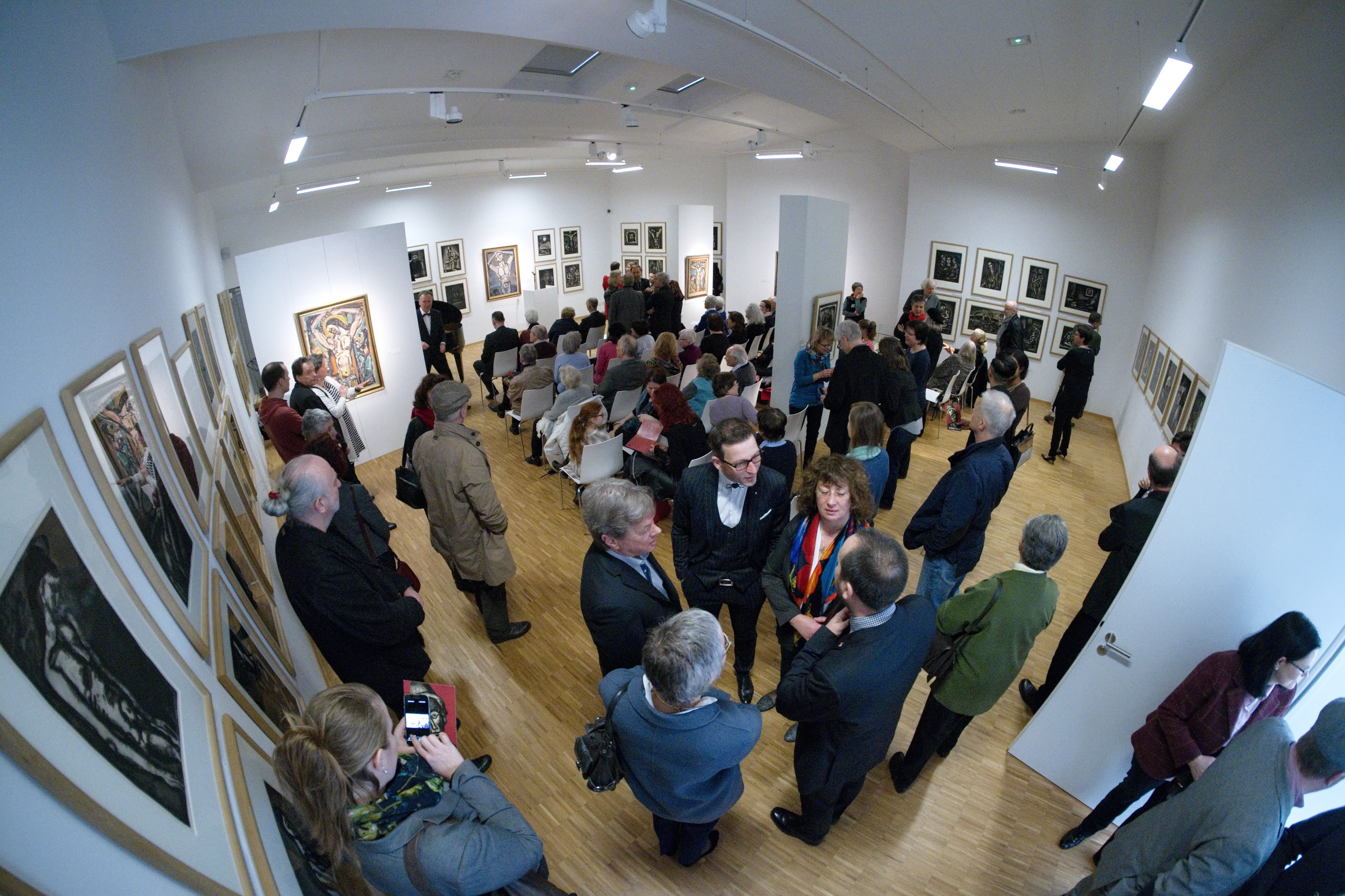 In einem Ausstellungsraum stehen und sitzen viele Menschen. Sie unterhalten sich oder schauen sich die Druckgraphiken an den Wänden an.