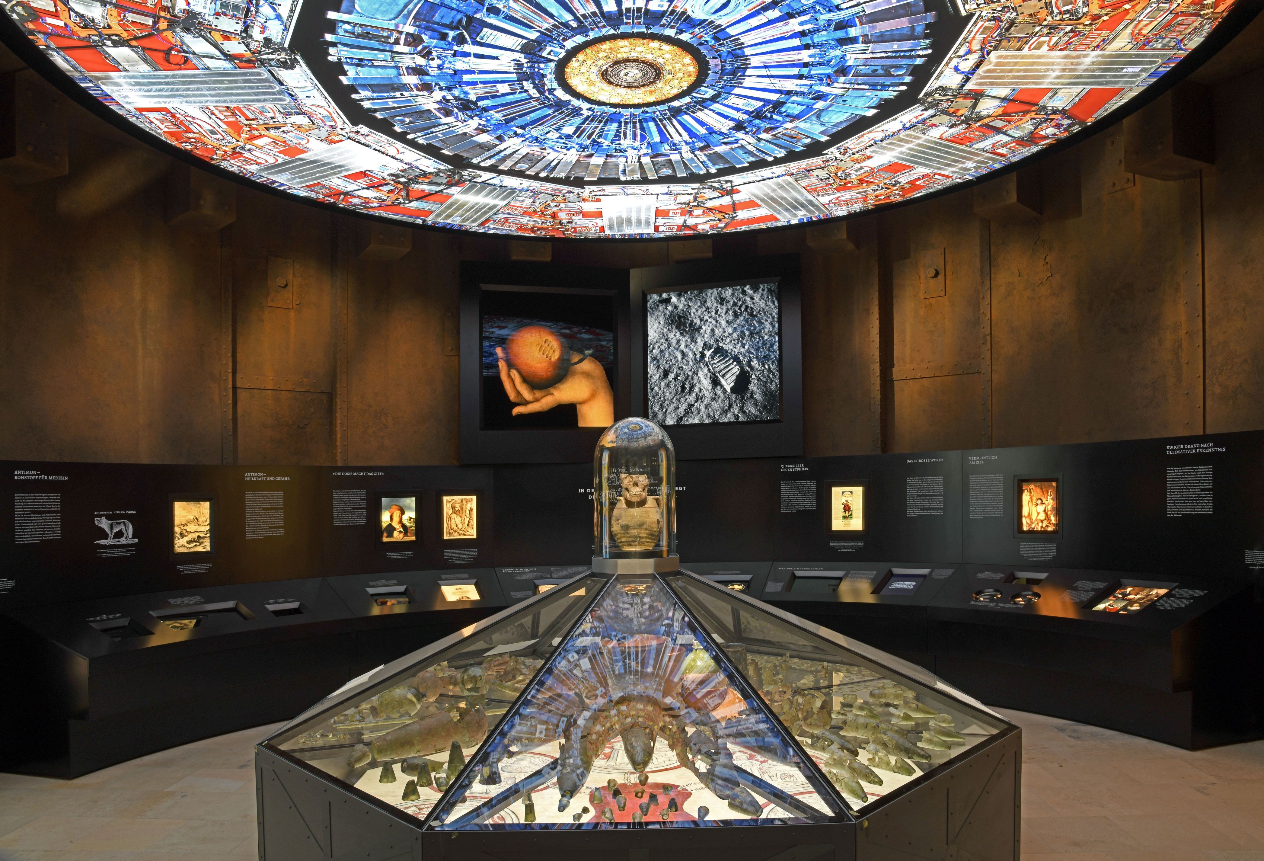 In der Mitte des Raumes steht eine achteckige Vitrine mit Funden aus der Alchemistenwerkstatt Wittenberg. Zahlreiche Gefäße, Scherben, Destillierkolben und Retorten gehören zu den Funden. Über der Vitrine hängt eine blau und rot leuchtende Kuppel. Im Hintergrund befinden sich weitere schwarze Vitrinen an der Wand. Darüber hängt das Bilder einer Hand, die einen Apfel hält und eines das Quecksilber darstellt.