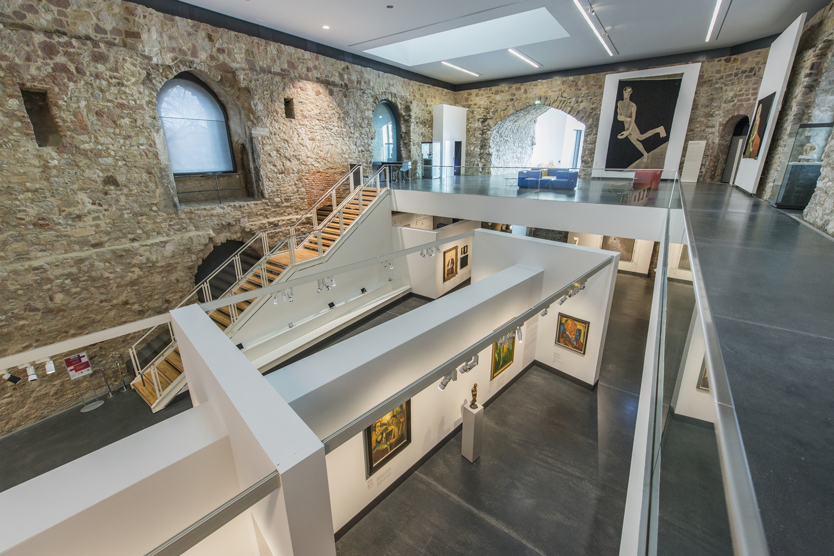 Blick in die Dauerausstellung "Wege der Moderne". Von einer Empore ist eine weiße freistehende Wand im raum zu sehen, an der die Gemälde angebracht wurde. Im Hintergrund ist eine Treppe zu sehen die auf die Empore führt. Ein großer Teppich hängt an der Wand.
