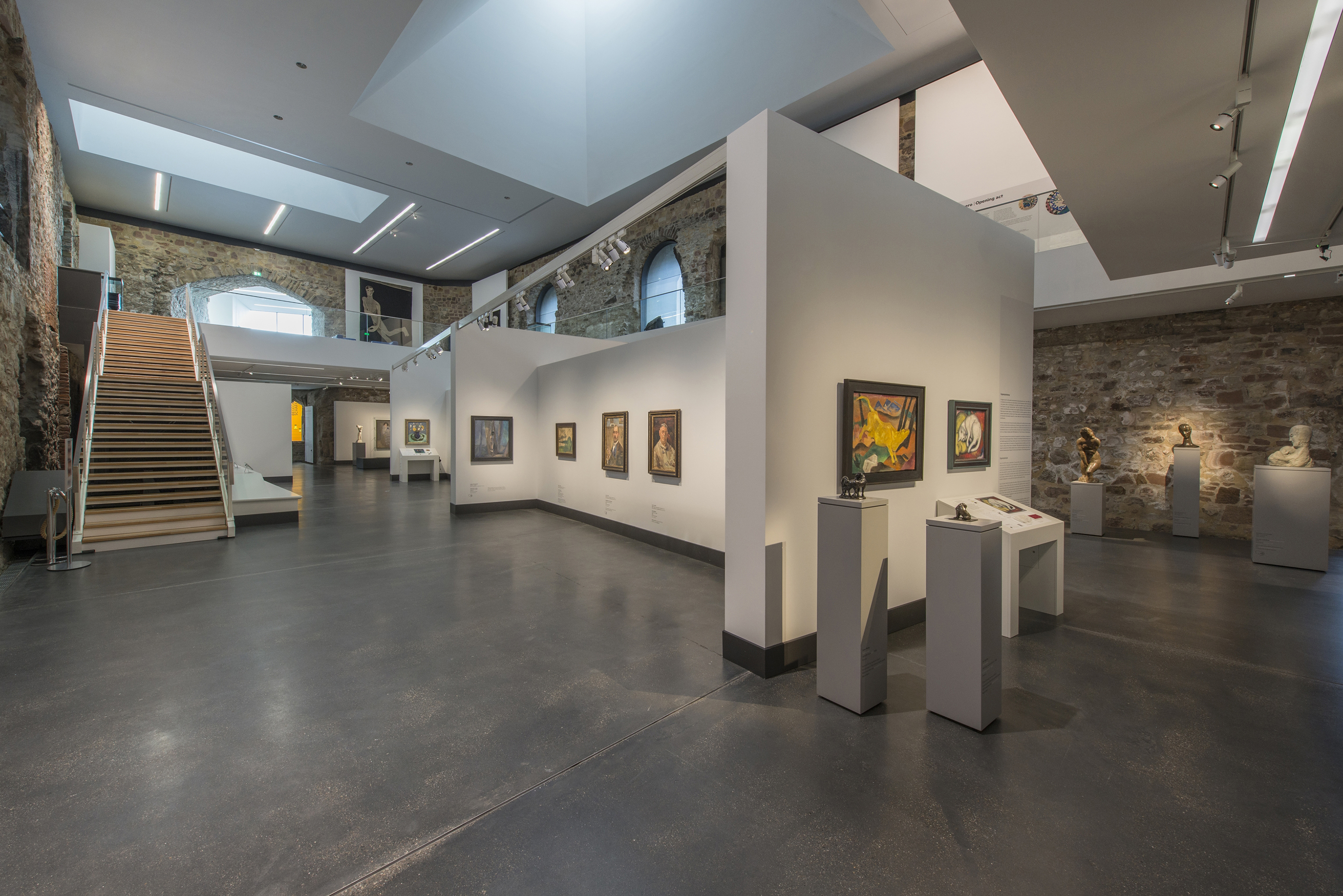 Blick in die Dauerausstellung "Wege der Moderne". An einer weißen Wand mitten im Raum hängen die Gemälde der Künstler der Moderne. Auf schmalen Stelen werden kleine und große Skulpturen präsentiert. Eine Treppe auf der linken Seite führt auf eine Empore.