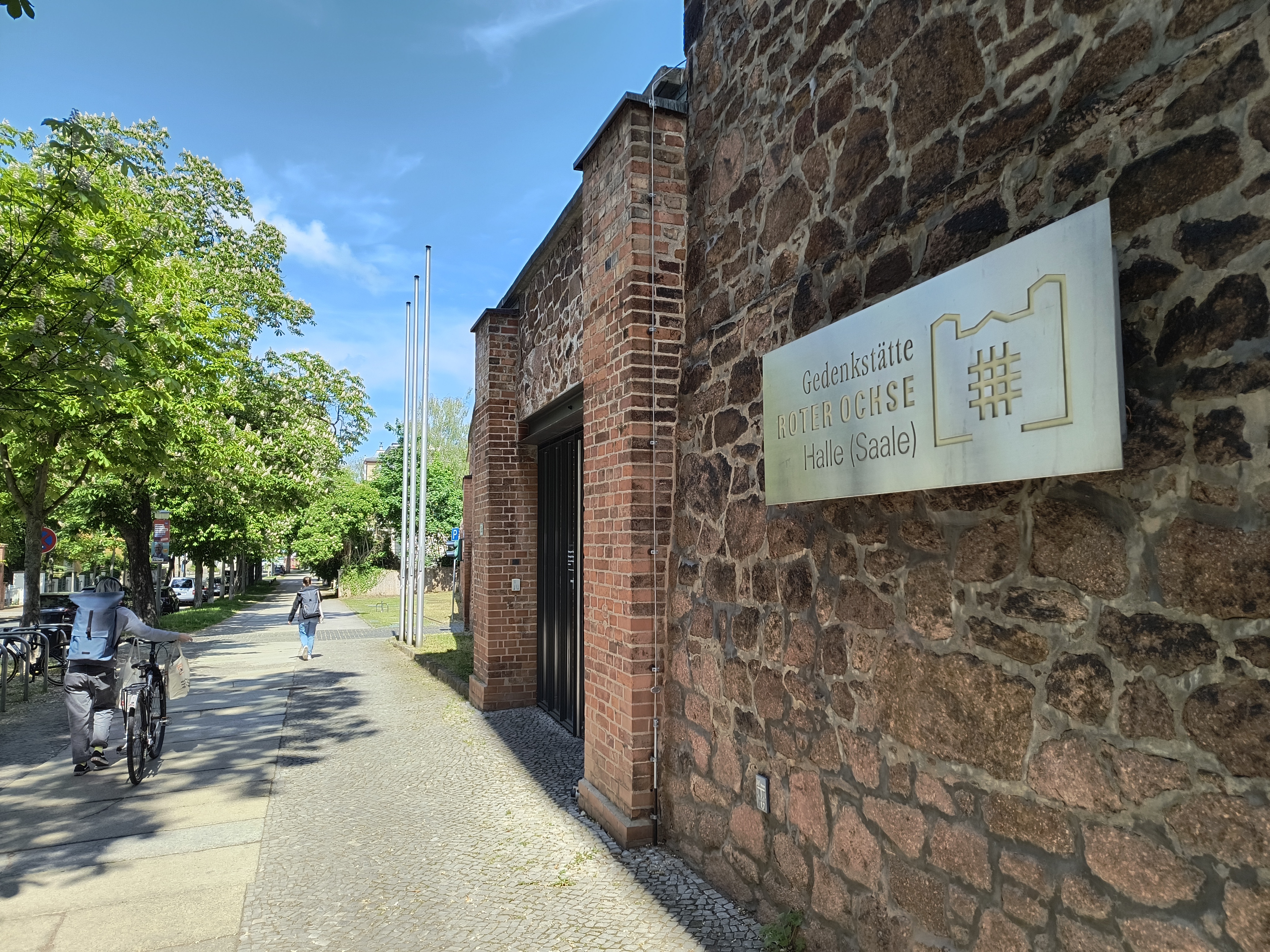 Auf der rechten Seite befindet sich eine rote Außenmauer mit einem metallischen Schild mit dem Verweis "Gedenkstätte roter Ochse Halle (Saale)". Dahinter befindet sich der Eingang zum Museum. Vor der Mauer befindet sich der Gehweg, auf dem eine Person ein Fahrrad schiebt und eine andere entlang läuft.