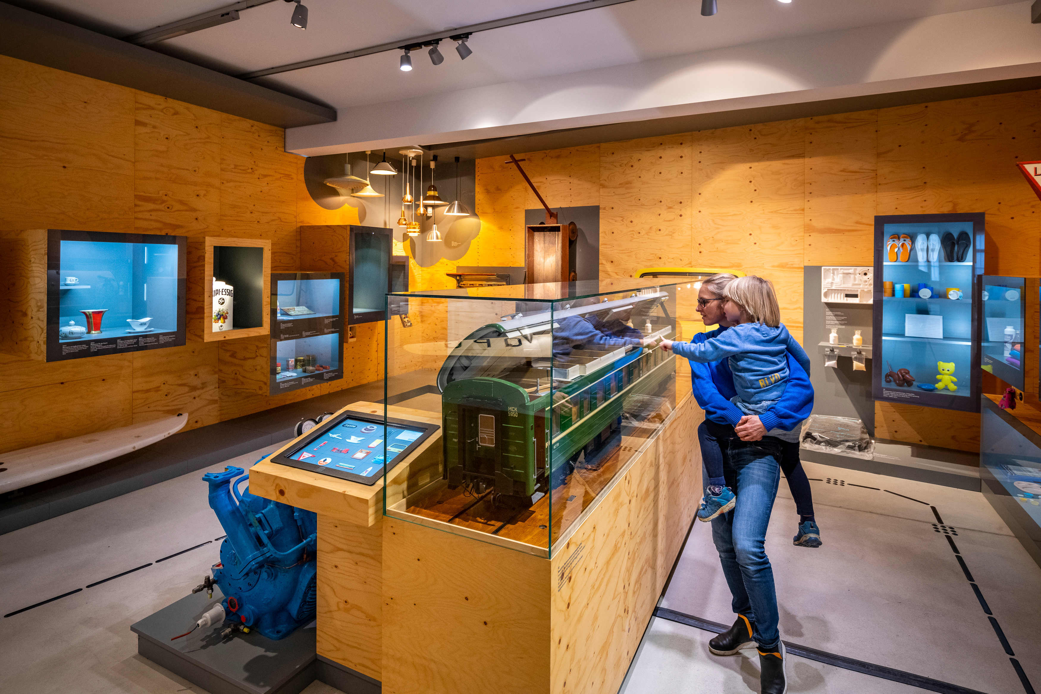 Vor einer Vitrine steht eine Mutter mit ihrem Kind im Arm. Sie betrachten das Modell eines grünen Zuges. Die Ausstellung zeigt verschiedene Alltagsobjekte, wie Vitrinen mit Schuhen, Bechern, Porzellangeschirr, Sielzeug und Lampen.