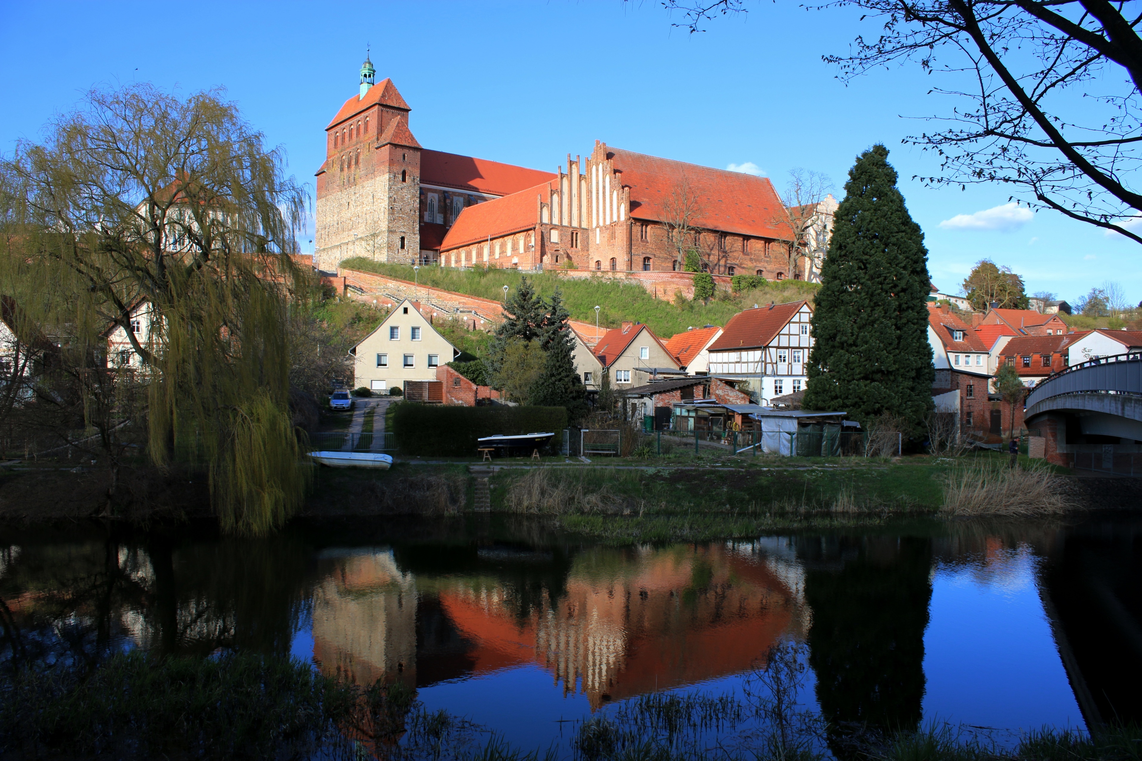 Der Havelberger Dom auf dem Hügel. Der Backsteinbau spiegelt sich im Wasser des Flusses vor dem Hügel wider. Am Fuße des Domes liegen Familienhäuser.