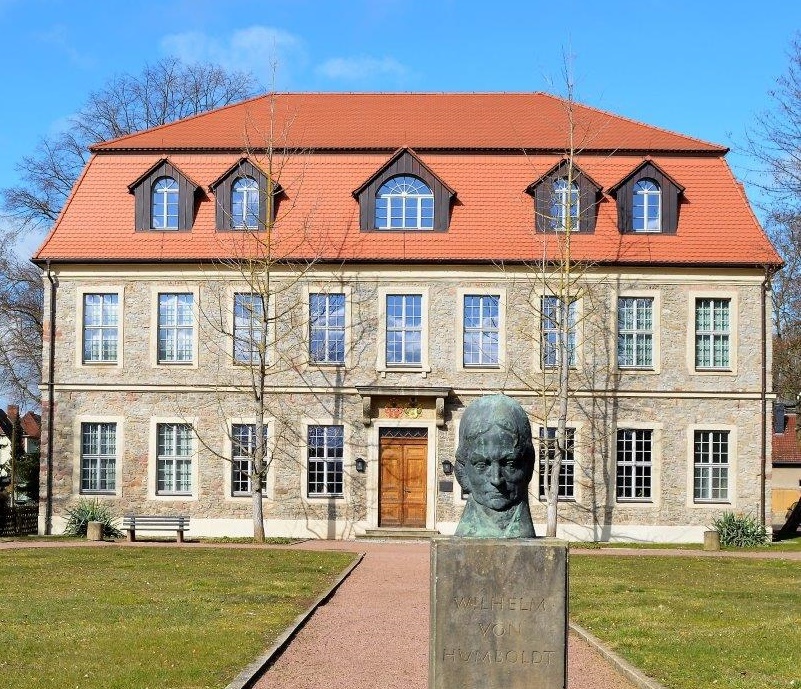 Das Humboldt-Schloss von Außen. Das Schloss besitzt eine graue Fassade mit Fenstern in zwei Reihen. Das Mansardenwalmdach ist rot. Vor dem schloss steht eine Büste auf einem Sockel mit der Inschrift Wilhelm von Humboldt.