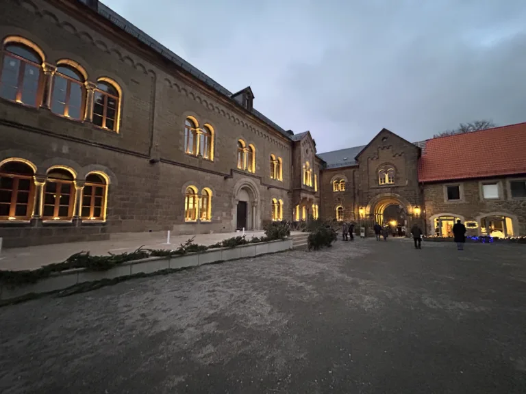 Das Bild zeigt den Innenhof des Crola Museums Ilsenburg. Die rundbogigen Fenster werden im Dämmerlicht beleuchtet und damit umrahmt. Zwischen den Fenstern stehen kleine Säulen. Durch ein rundbogiges Stufenportal kommen Besucher*innen in den Hof.