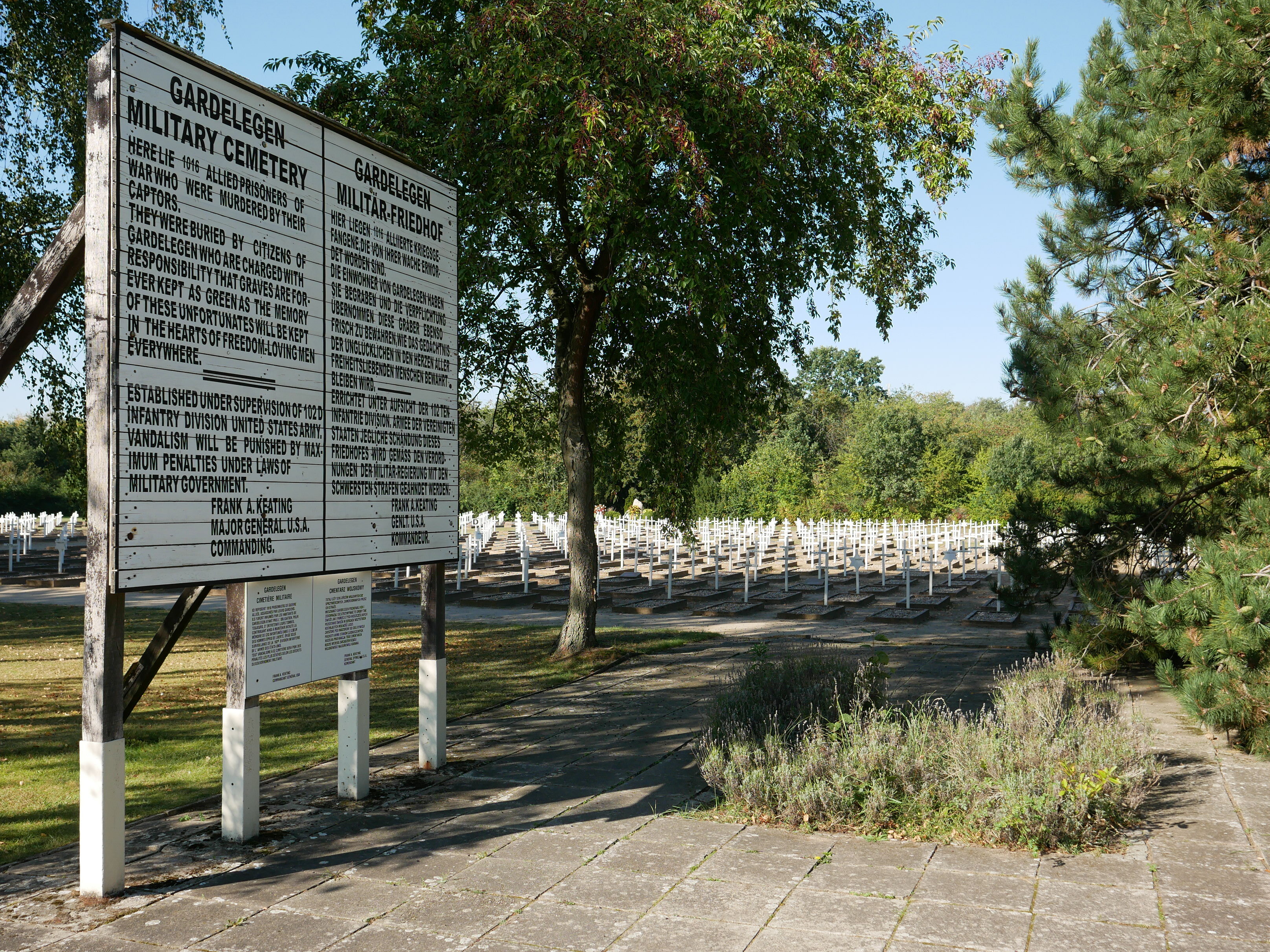 Der Ehrenfriedhof für die Ermordeten des Massakers von Gardelegen. Eine große weiße Gedenktafel erinnert an die ermordeten Alliierten. Im Hintergrund sind die Gräber und weiße Kreuze zwischen Bäumen zu sehen.