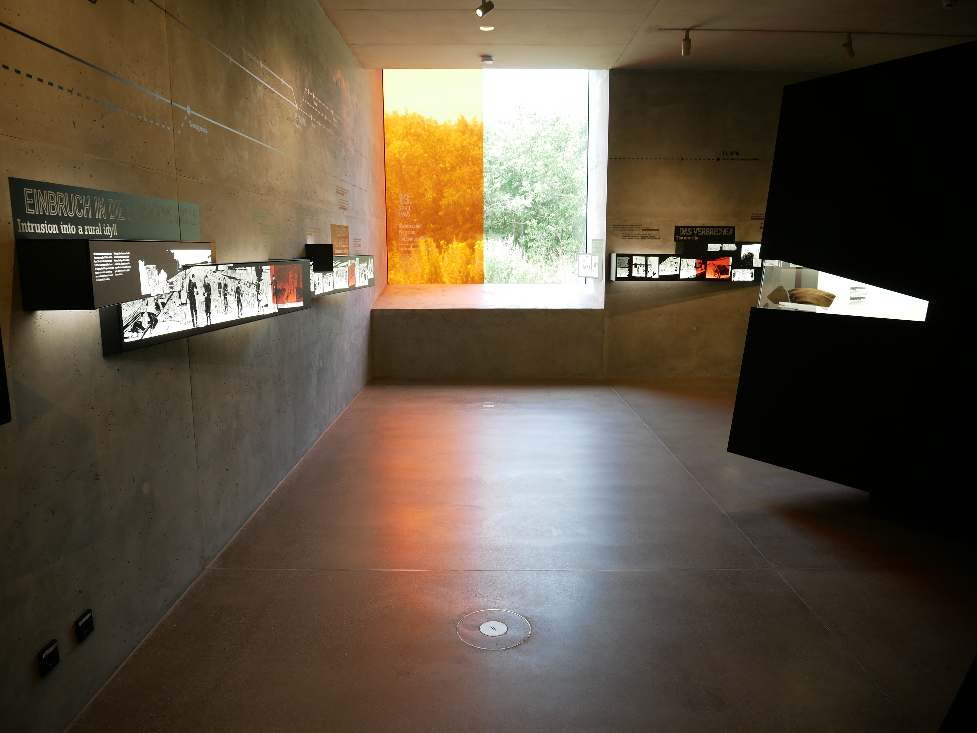 Ausstellung "Gardelegen 1945. Das Massaker und seine Nachwirkungen". In einem dunklen Raum mit Betonwänden hängen mehrere lange schwarze Kästen mit beleuchteten Fotografien der Zeit um das Massaker. Eine weitere Vitrine zeigt eine militärische Kopfbedeckung. In den Raum fällt Licht durch ein großes quadratisches Fenster, das zur Hälfte mit einer orangenen Folie abgeklebt wurde.