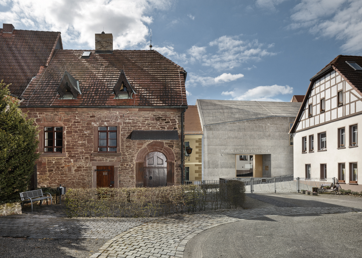 Zwischen einem roten Steinhaus und einem weißen Fachwerkhaus zeigt das graue Museumsgebäude des Elternhauses von Luther hervor. Ein großer Eingang führt in das Innere des Museums.