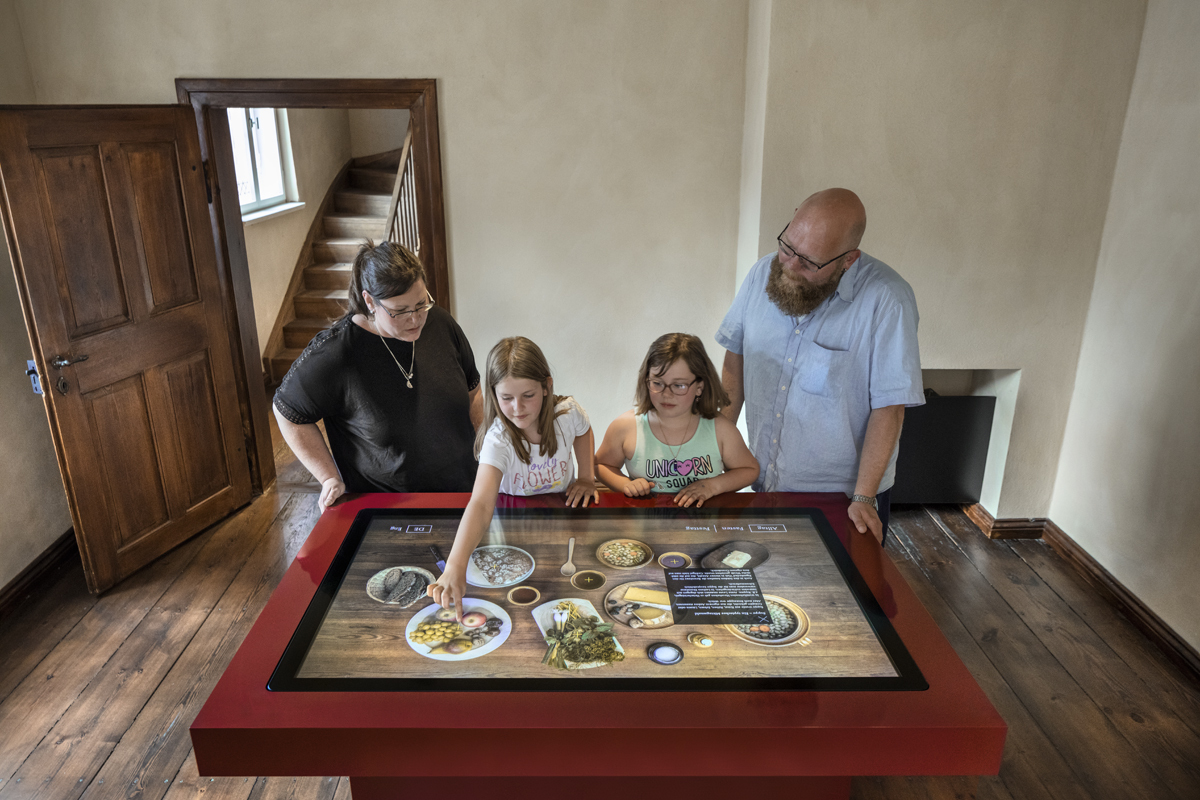 Eine Familie mit zwei Kindern steht vor einem Tisch mit einem Touchscreen. Sie ordnen darauf Bilder von Lebensmitteln an.