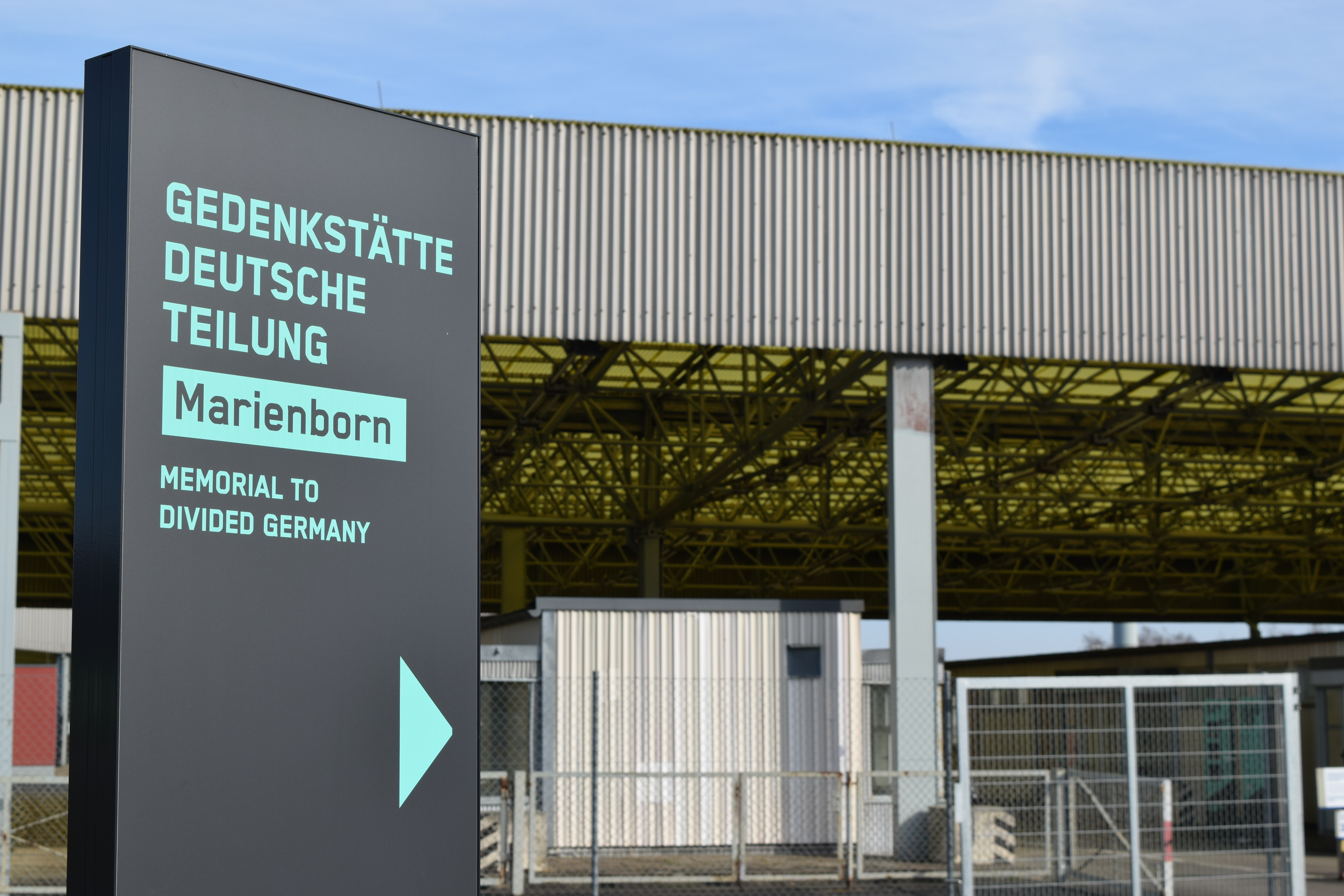 Der Eingangsbereich der Gedenkstätte. Eine Tafel im Vordergrund verweist auf die Gedenkstätte deutsche Teilung Marienborn. Im Hintergrund ist ein mit grauem Wellblech verkleidetes Gebäude zu sehen.