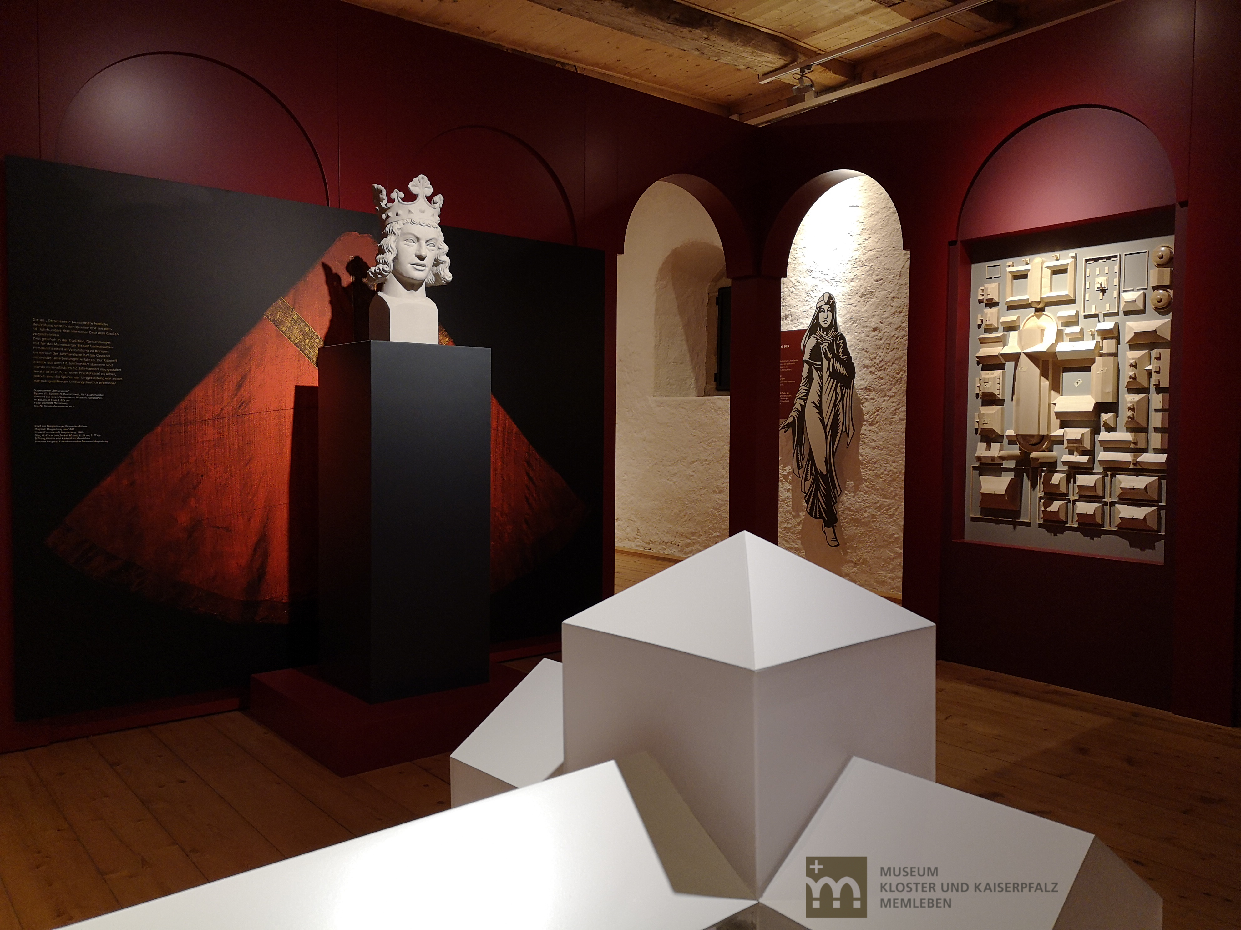 Hier wird die Ausstellung "Wissen und Macht. Der heilige Benedikt und die Ottonen" gezeigt. Im Vordergrund ist ein Teil eines Architekturmodells zu sehen. Im Hintergrund steht die gekrönte Büste von Otto dem Großen, dahinter ist eine rote Kasel auf einer Tafel dargestellt. Ein weiteres Modell hängt an der nebenliegenden Wand.