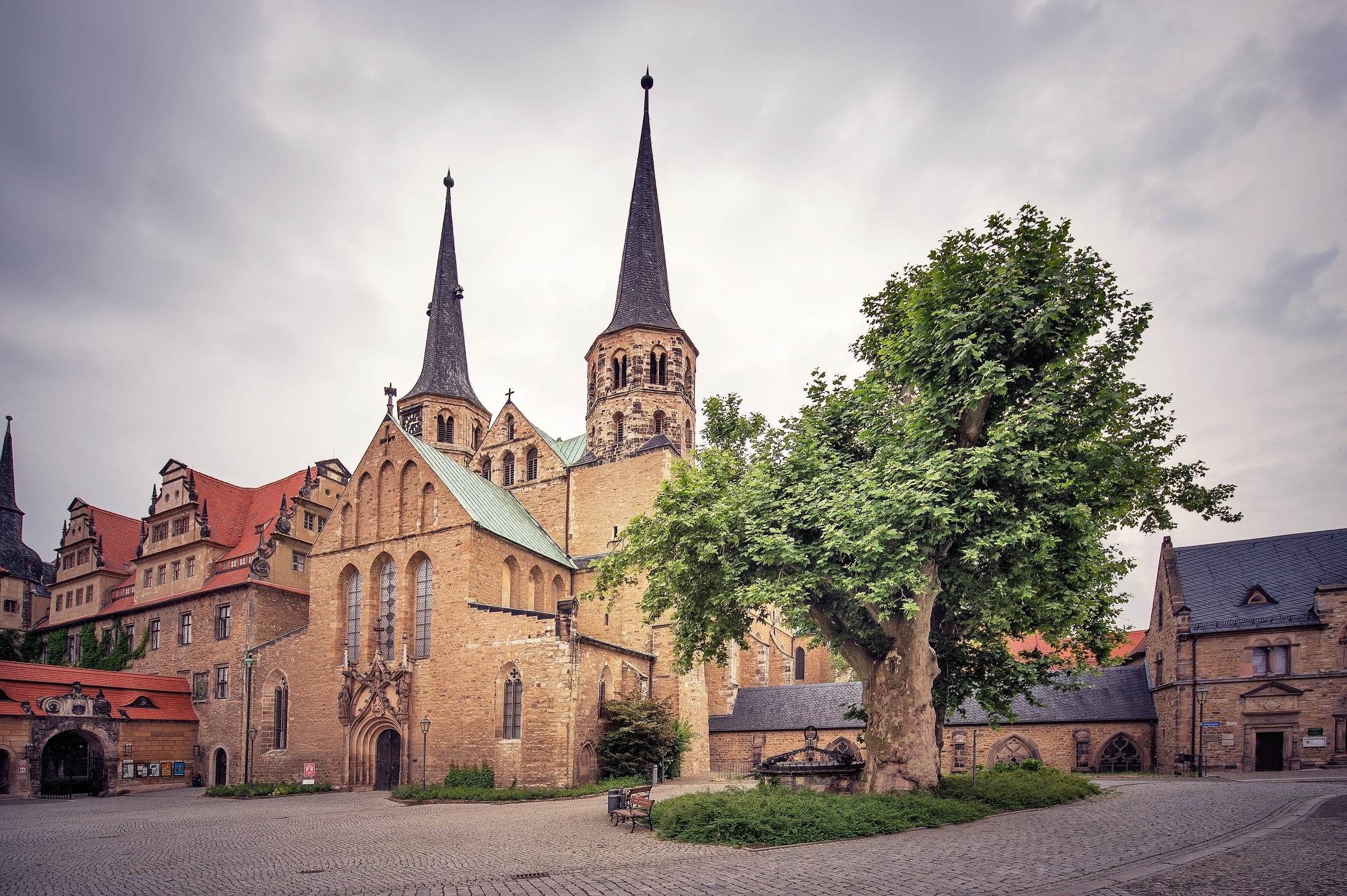 Außenansicht Kaiserdom Merseburg. Auf dem Platz vor dem Dom steht ein großer Baum. Dahinter befinden sich Kapitelhaus, Kreuzgang und Dom. Vor den westlichen Türmen ist die Vorhalle mit einem ausgeschmückten Portal erkennbar. Daneben befindet sich das Schloss.