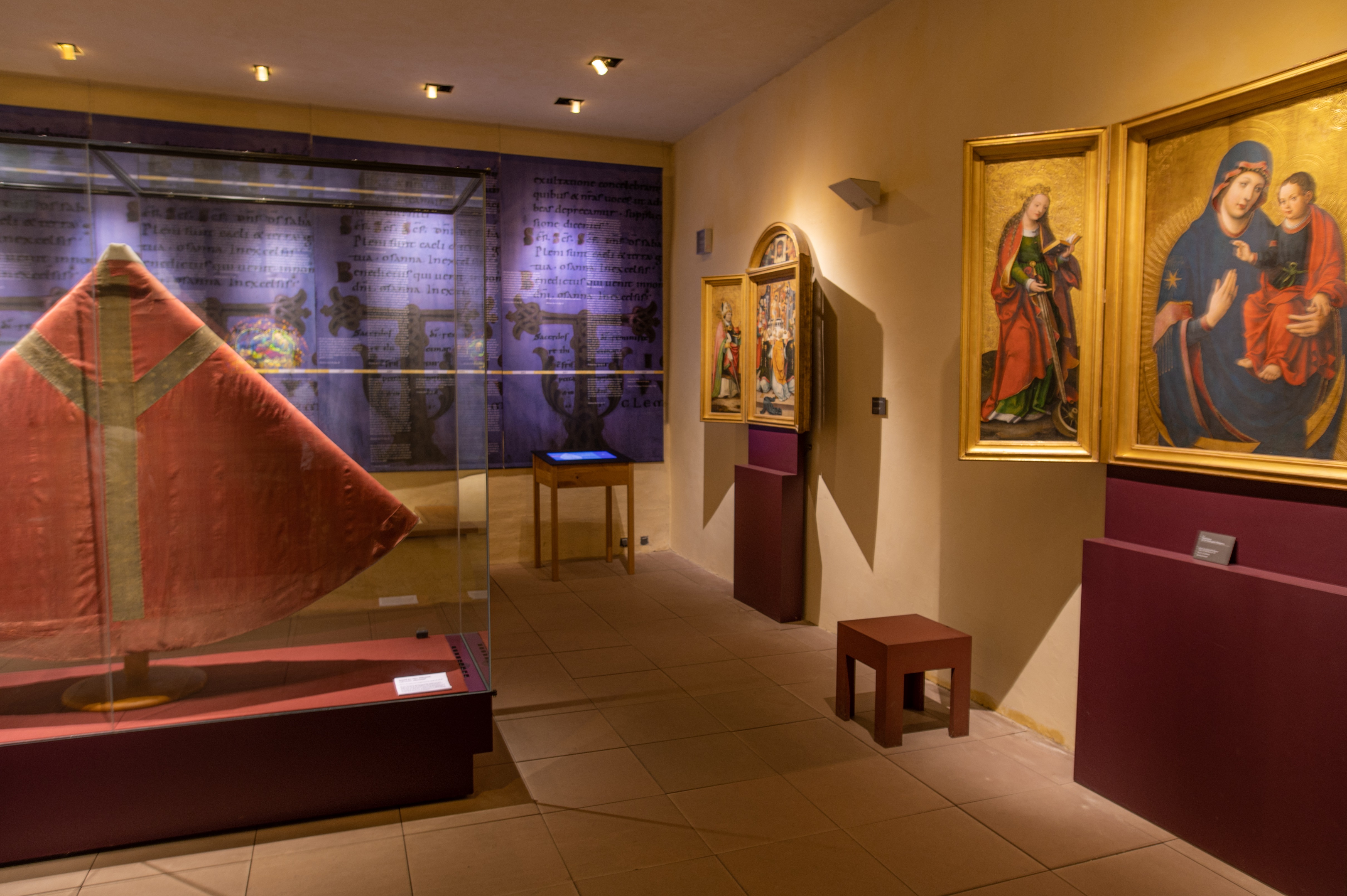 Im Kapitelhaus des Merseburger Domes befindet sich in der Mitte des Raumes ein rotes Kirchengewand (Pluviale). An den Wänden repräsentieren Altäre verschiedene Heilige und deren Geschichten. Im Hintergrund hängen lila Stoffe mit Beschriftungen.