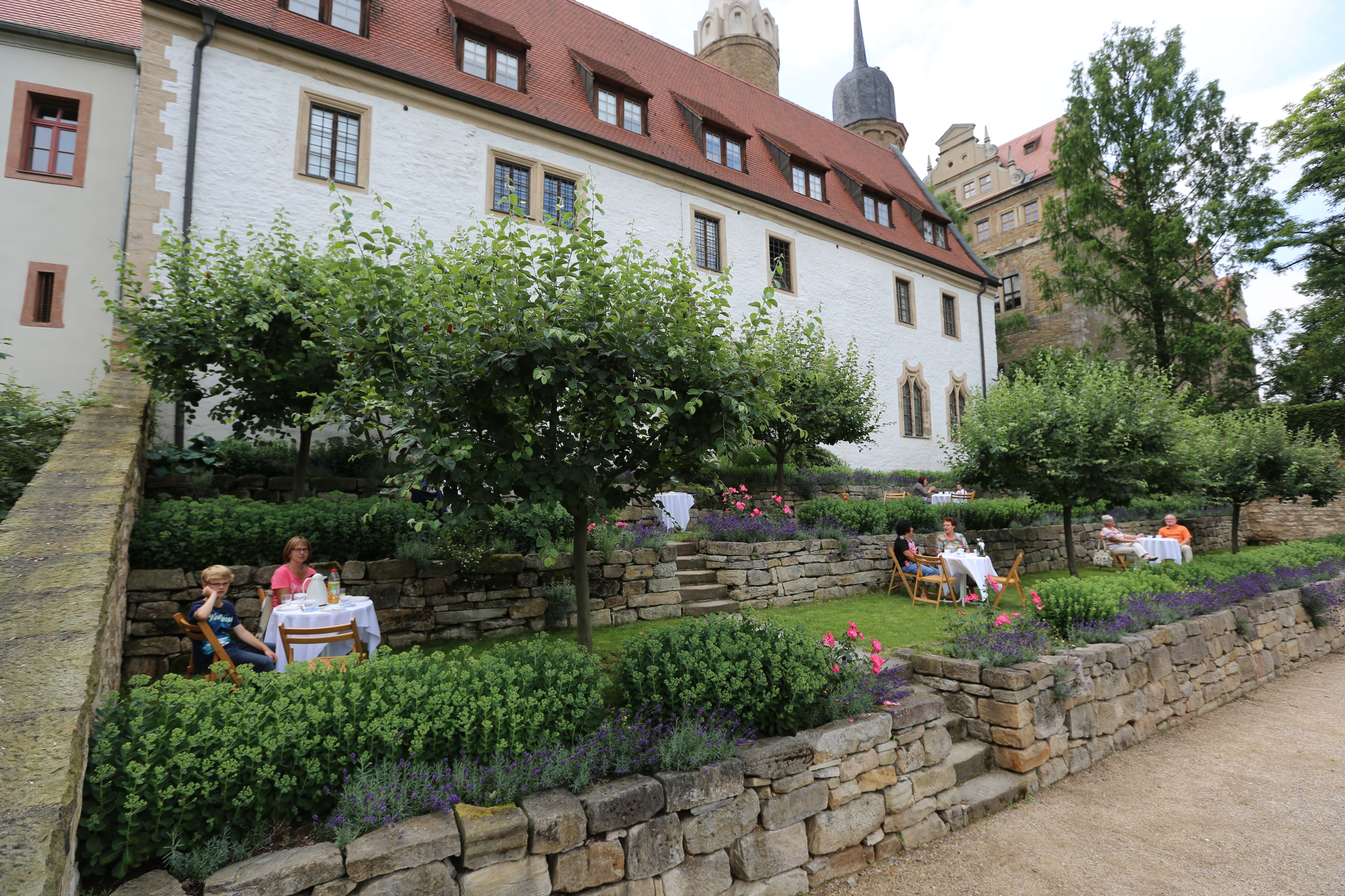 Kapitelhausgarten des Merseburger Domes. Hinter dem Kapitelhaus befindet sich ein arten auf zwei Ebenen. In den Beeten wachsen Rosen und Lavendel und kleine Bäume spenden Schatten. Auf der Wiese sitzen Menschen an Tischen und trinken Kaffee.