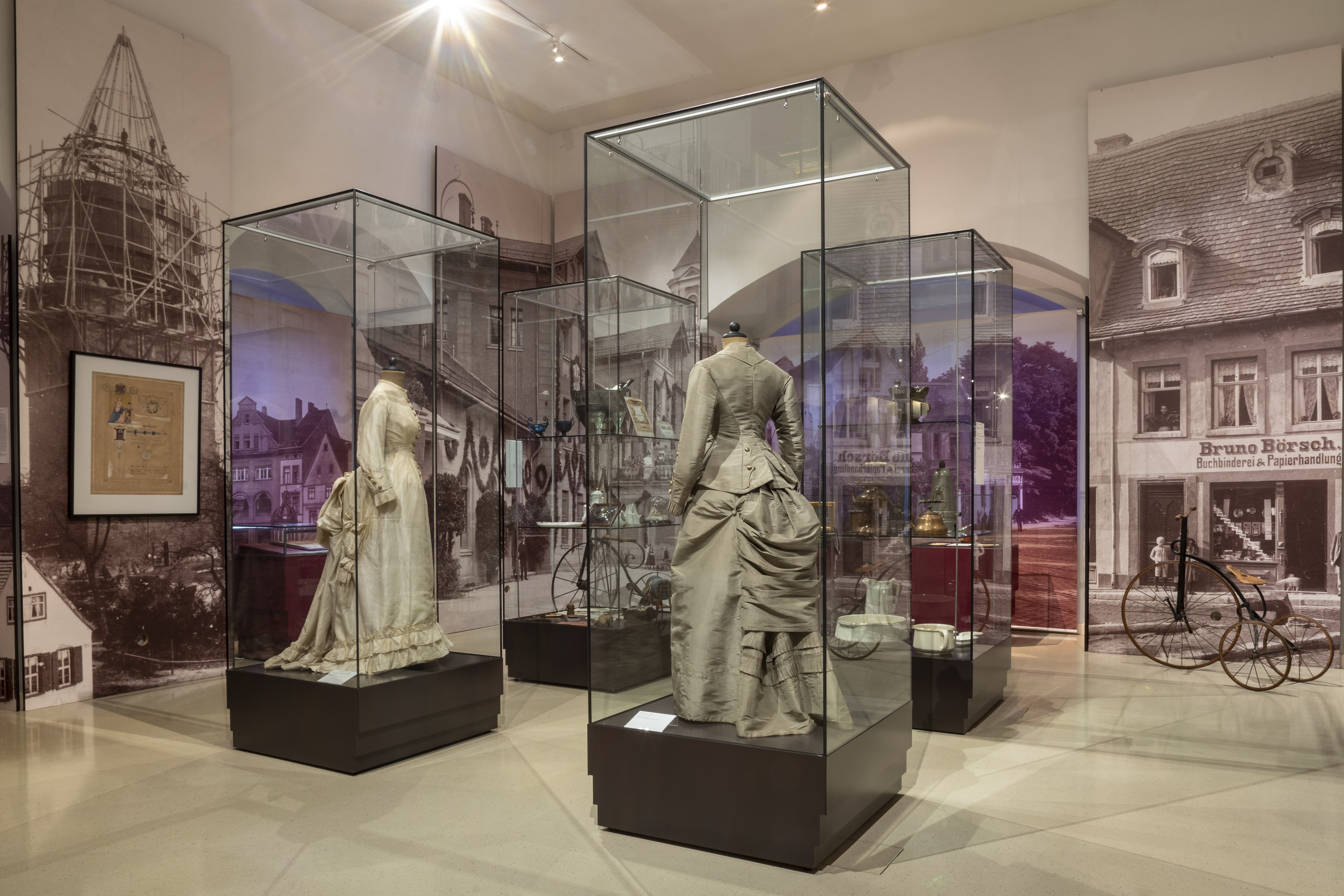 Merseburg zur Gründerzeit. In hohen Vitrinen werden die Kleidermode und Alltagsgegenstände während der Gründerzeit präsentiert. An den Wänden zeigen Aufnahmen das historische Merseburg.