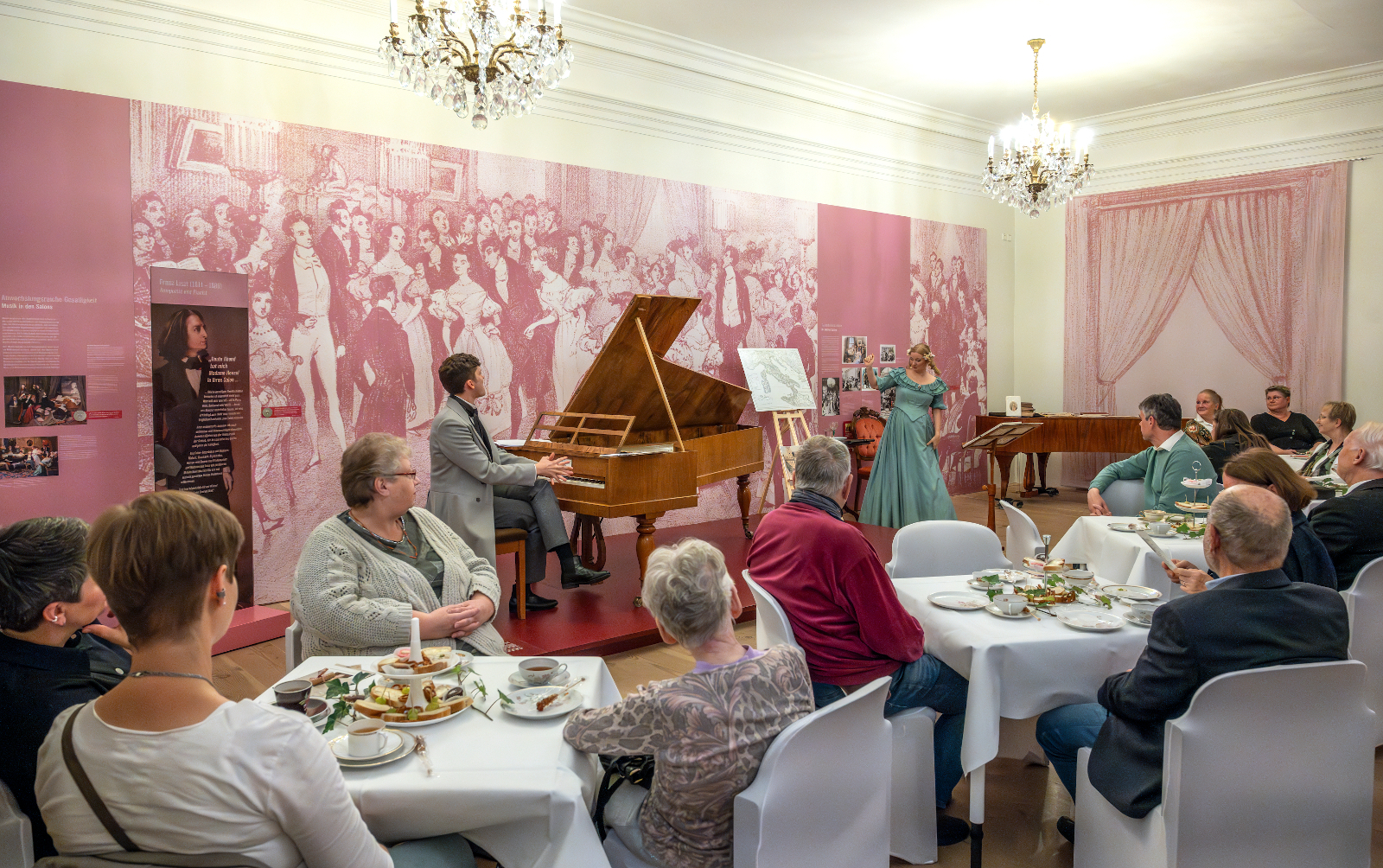 Lebendige Musiktradition im historischen Ambiente: Ein klassisches Konzert gepaart mit einer Teezeremonie im Salon eines kulturellen Museums. Eine Gesellschaft sitzt an weißen gedeckten Tischen. Auf einer kleinen Bühne sitzt ein Mann an einem Holzflügel und eine Frau in einem blauen langen Kleid daneben singt.
