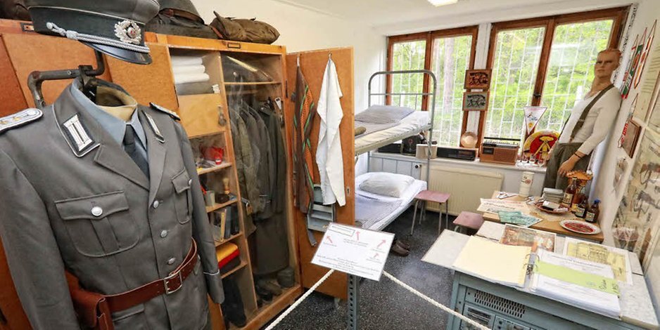In der NVA Stube wird das Zimmer eines Mitarbeiters gezeigt. Im Vordergrund wird eine Uniform präsentiert. Ein geöffneter Schrank dahinter, zeigt weitere Uniformen und Utensilien des Alltags. Ein Schreibtisch mit geöffneten Ordnern verweisen auf Unterlagen und die Arbeit in der Kaserne.