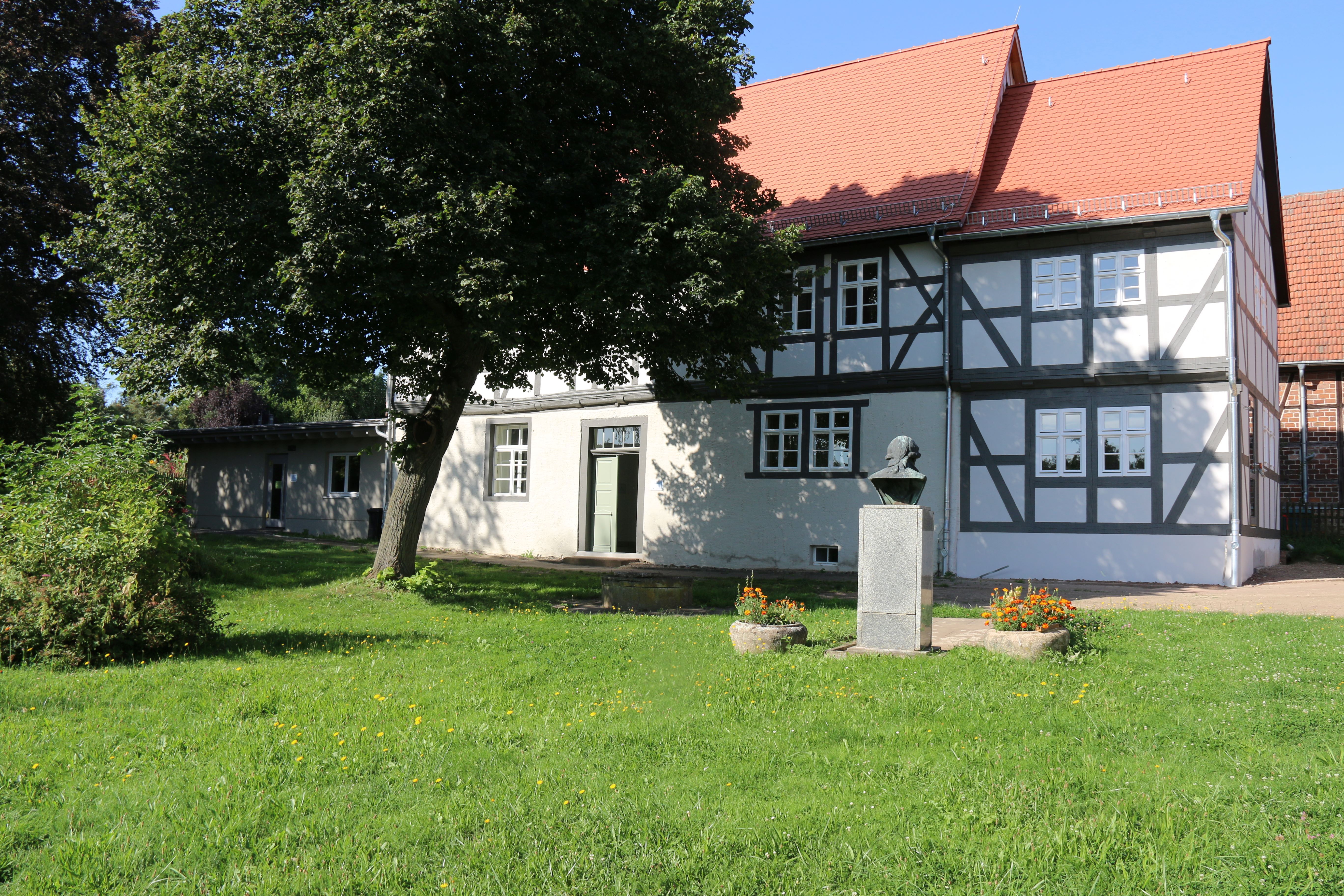 Das helle Museumsgebäude mit dunklem Fachwerk steht auf einer grünen Wiese. Davor befindet sich ein Sockel mit einer Büste.