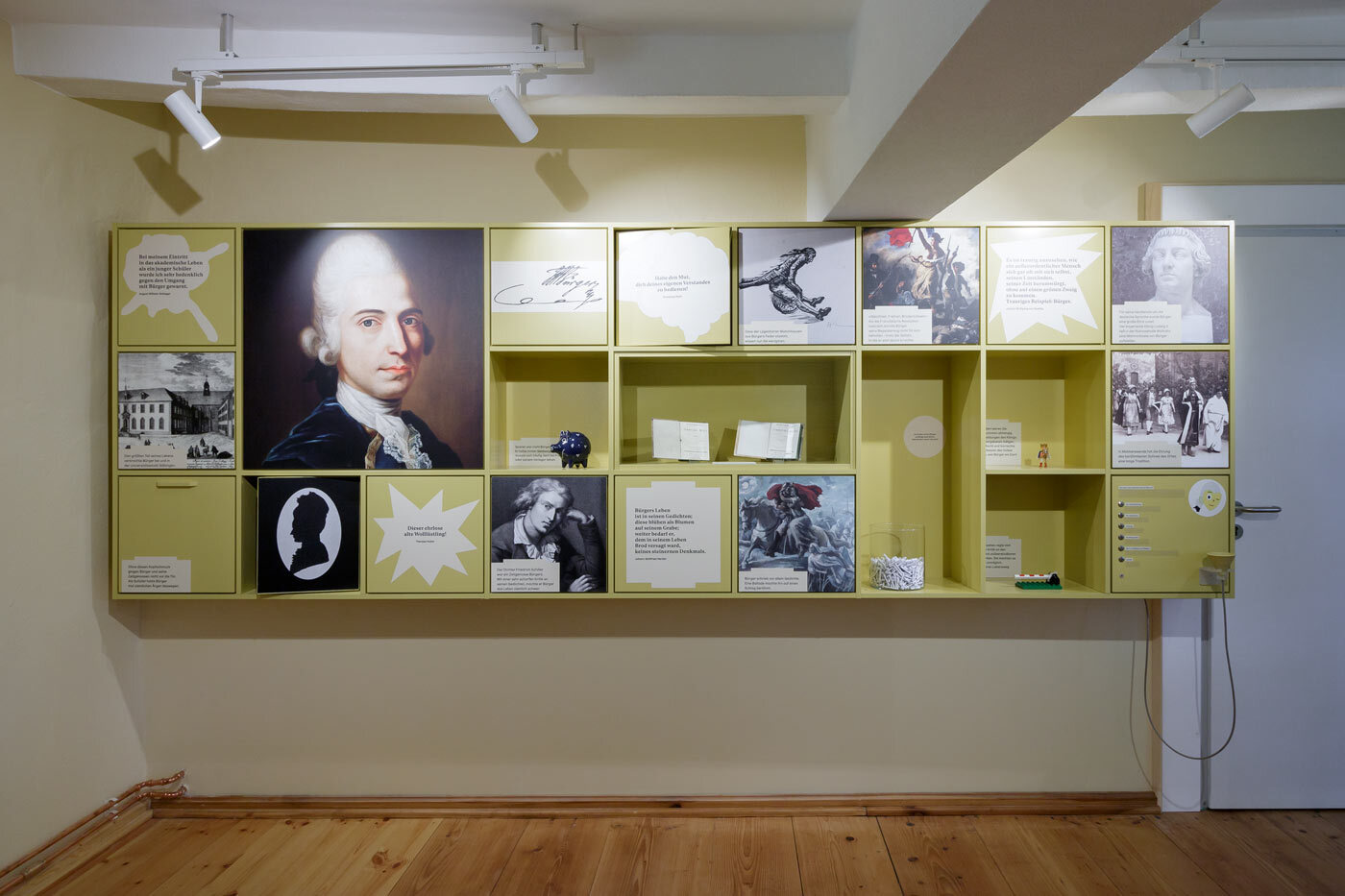 Eine gelbe Wandvorrichtung mit bebilderten Boxen dient als Prolog zur Ausstellung. Abbildungen von Porträts, Kupferstichen, Schlachtszenen, Scherenschnitten und Zitaten führen in die Zeit ein.