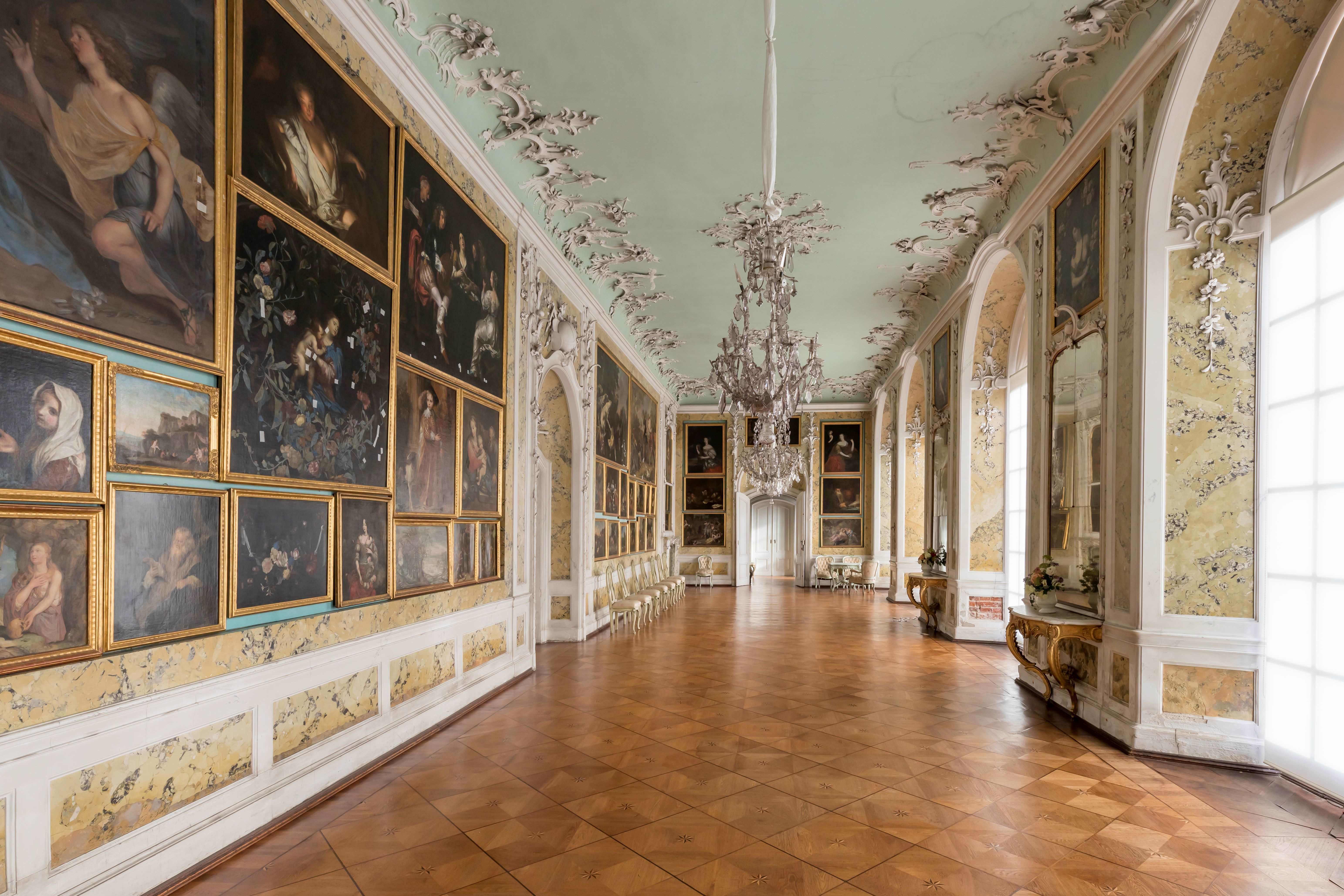 Der Galeriesaal im Schloss Mosigkau. Gegenüber der Galeriewand befinden sich große halbrunde Fenster. Dazwischen hängen große Spiegel. Die Decke ist geschmückt mit Rocaille Ornamenten.