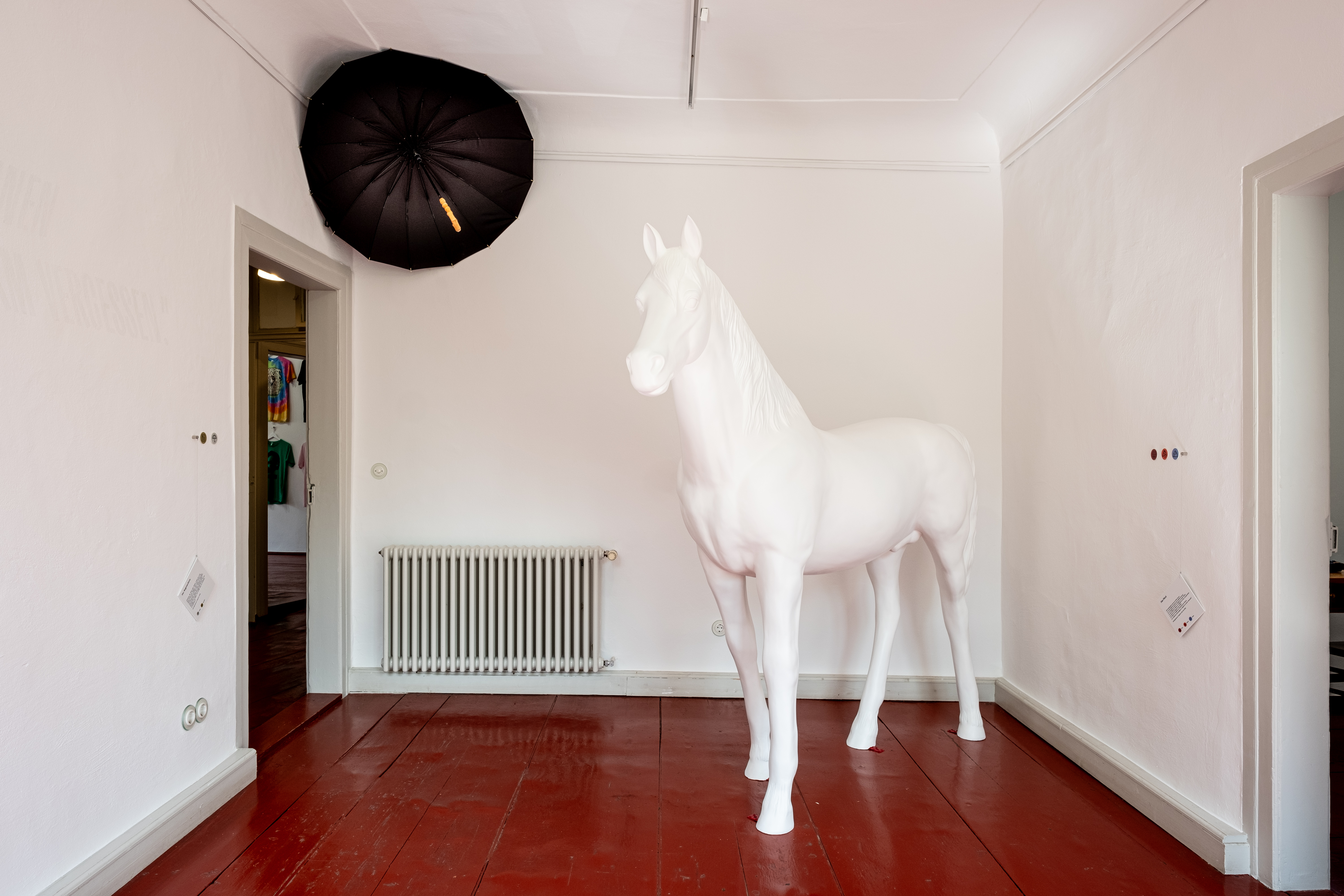 In Ausstellungsraum 3 hängt ein schwarzer Regenschirm in der linken oberen Ecke des Raumes. Die Figur eines weißen Pferdes steht mitten im Raum auf dem roten Fußboden.