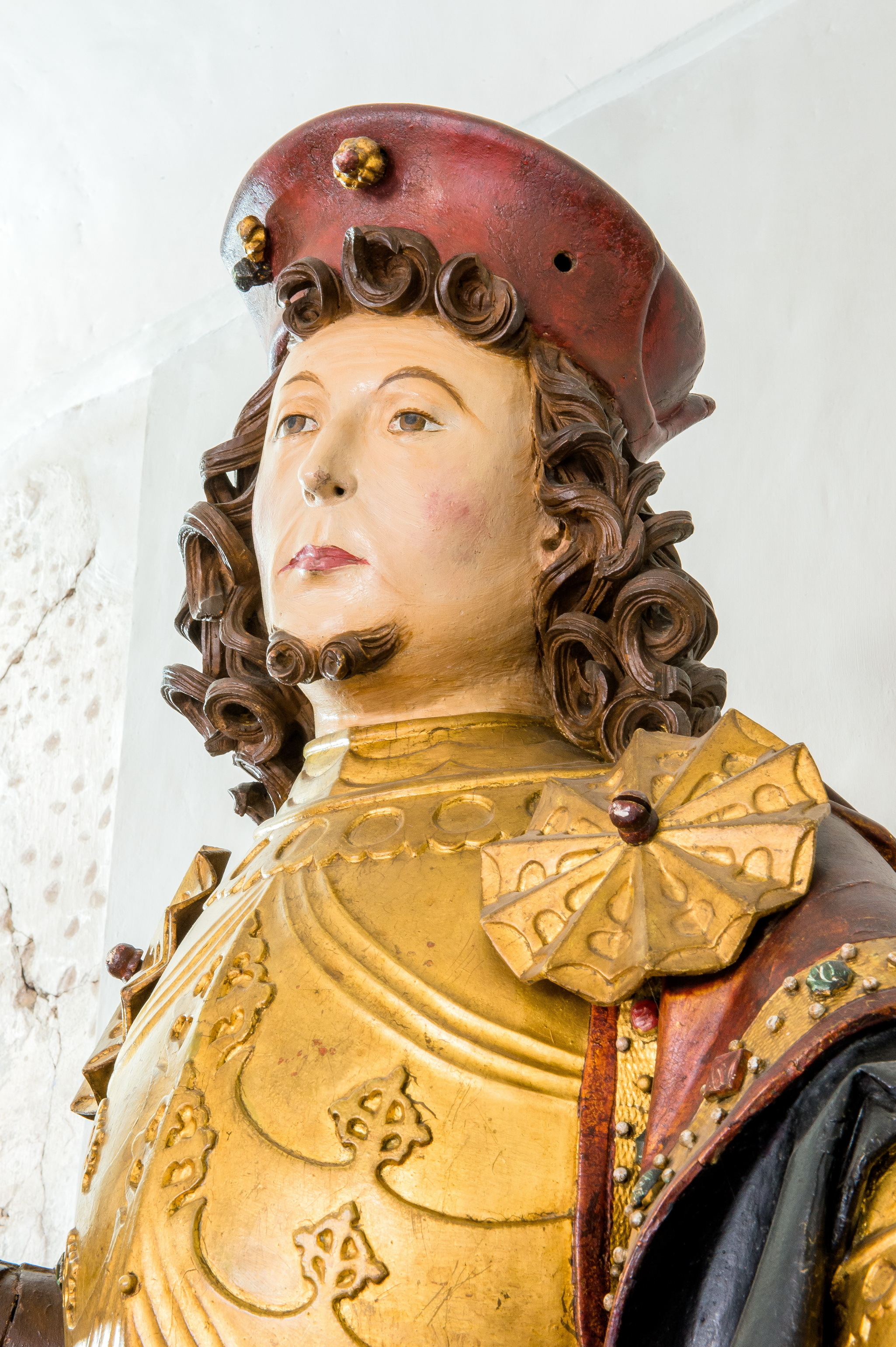 Skulptur des Heiligen Wenzel um 1530. Die Holzskulptur ist farbig gefasst. Sie trägt eine rote Kopfbedeckung, eine goldene Rüstung und einen roten Mantel. Braune Locken und ein Bart am Kinn rahmen das Gesicht ein.