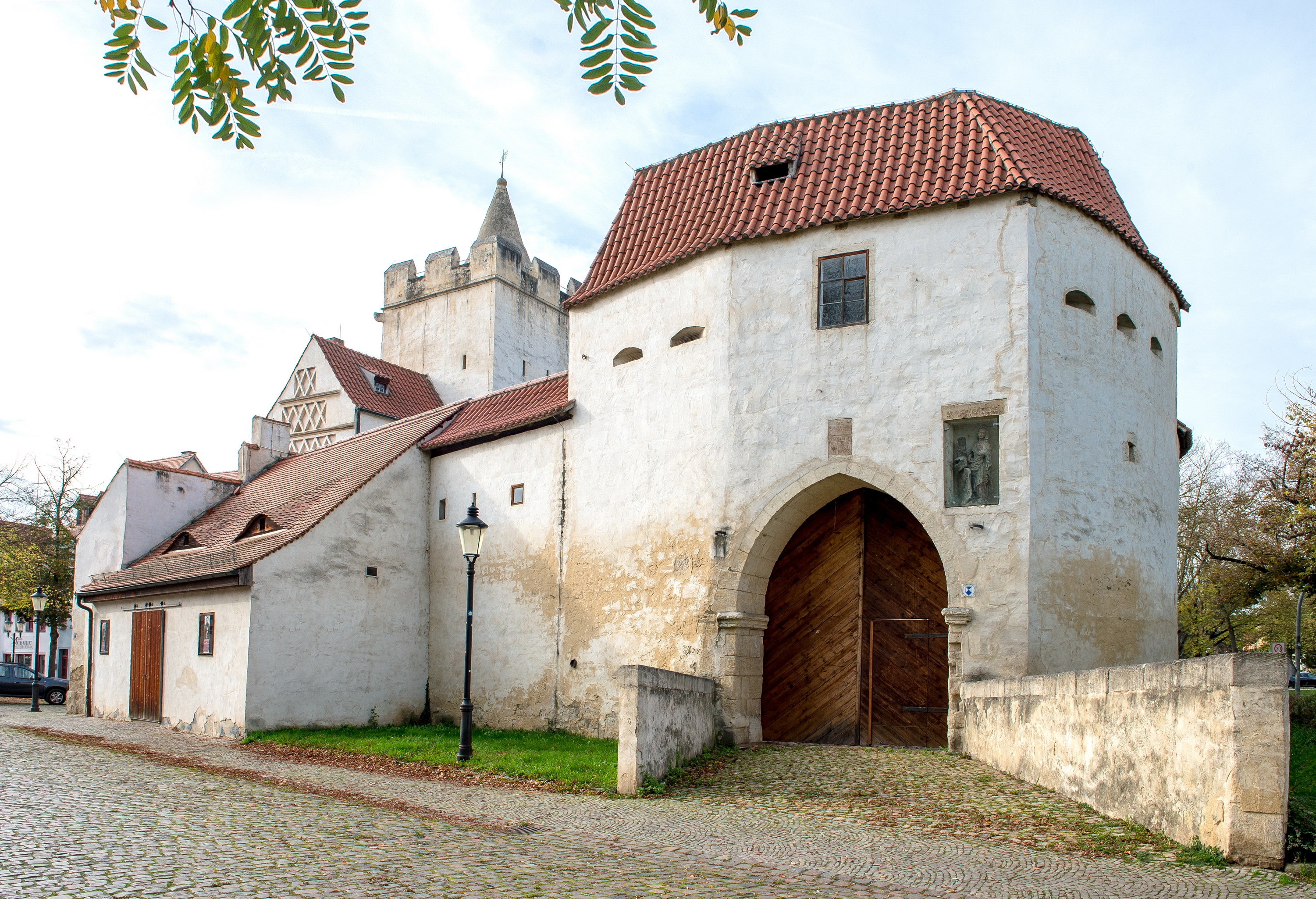 Die Toranlage besteht aus einer weißen Mauer mit einem roten Ziegeldach. Ein Holzportal ermöglicht den Zugang ins Innere. Im Hintergrund ist das angebaute Akzisehaus erkennbar.