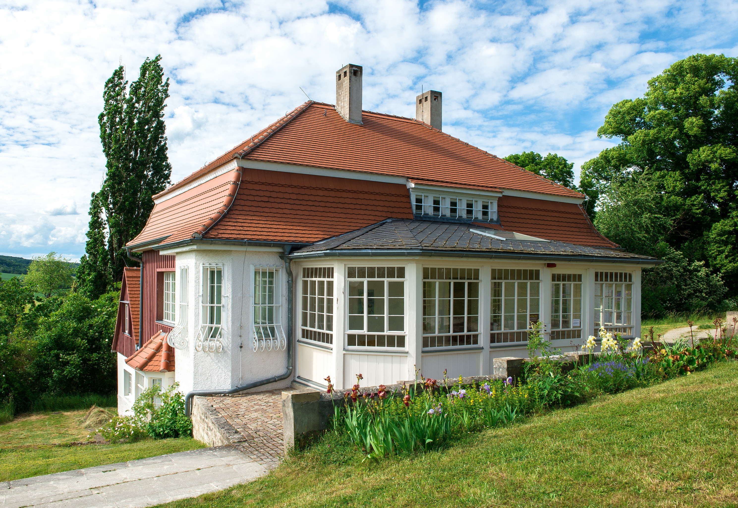 Das Max Klinger Haus besitzt eine weiße Fassade mit zahlreichen Fenstern und einem roten Mansardenwalmdach aus Ziegeln. Vor dem Besuchereingang wachsen bunte Blumen.