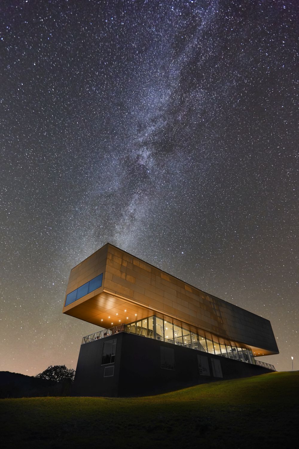 Das moderne kubische Gebäude des Besucherzentrums der Arche Nebra ist hell erleuchtet und aus von unten fotografiert wurden. Auf dem dunklen Nachthimmel dahinter ist die Milchstraße erkennbar.