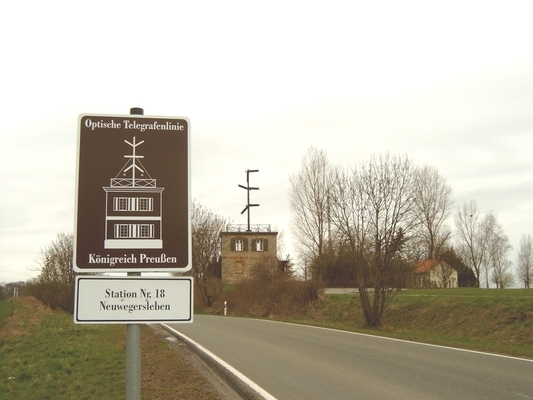An einer Straße steht ein braunes Schild mit dem Verweis: Optische Telegrafenlinie Königreich Preußen. Darunter befindet sich ein weißes kleines Schild mit folgender Aufschrift: Station Nr. 18 Neuwegersleben. Im Hintergrund ist das kleine Gebäude des Telegrafiemuseums erkennbar.