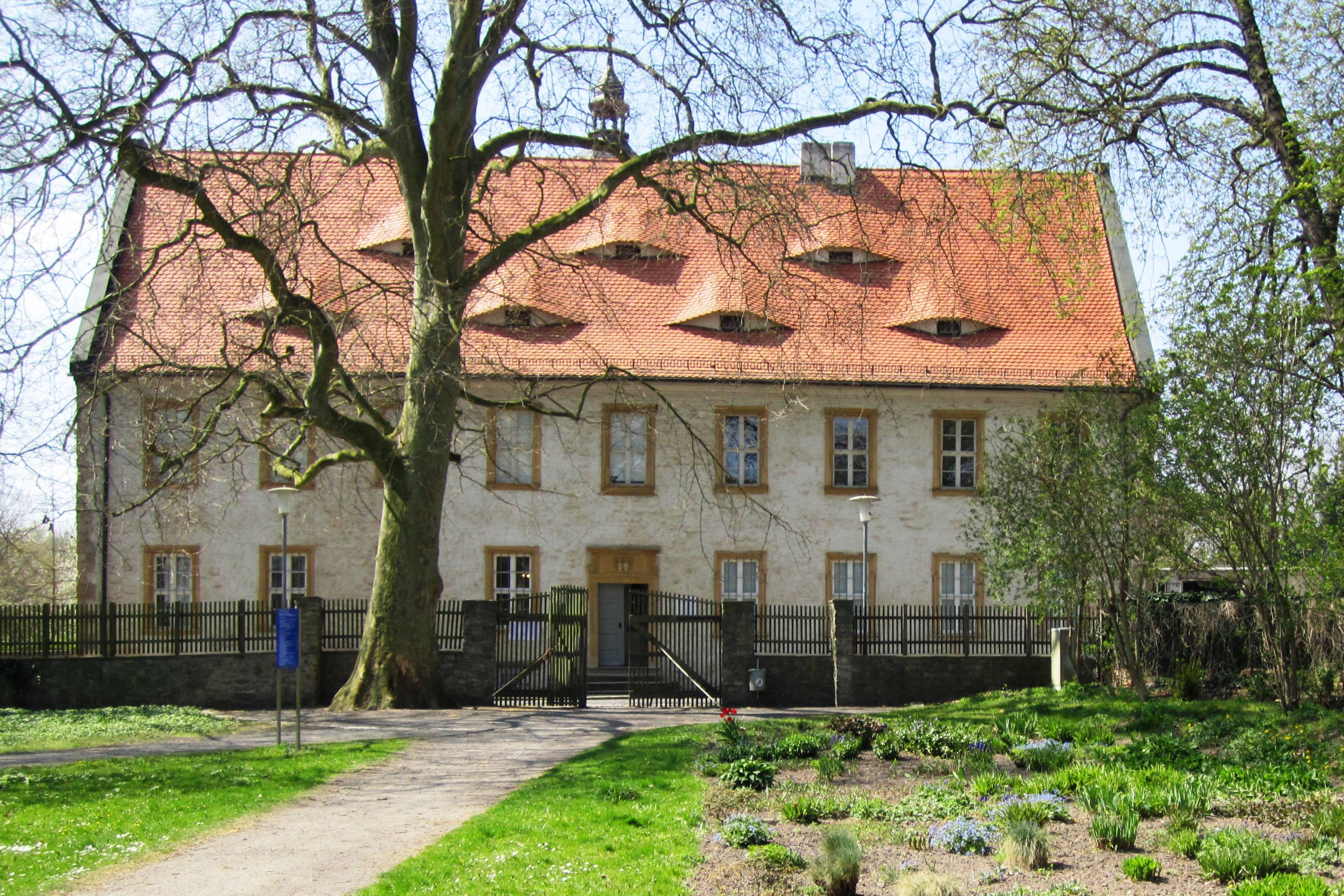 Novalis Schloss Seitenansicht. Vor einer hellen Hausfassade liegen Beete in eine Parkanlage eingebettet.