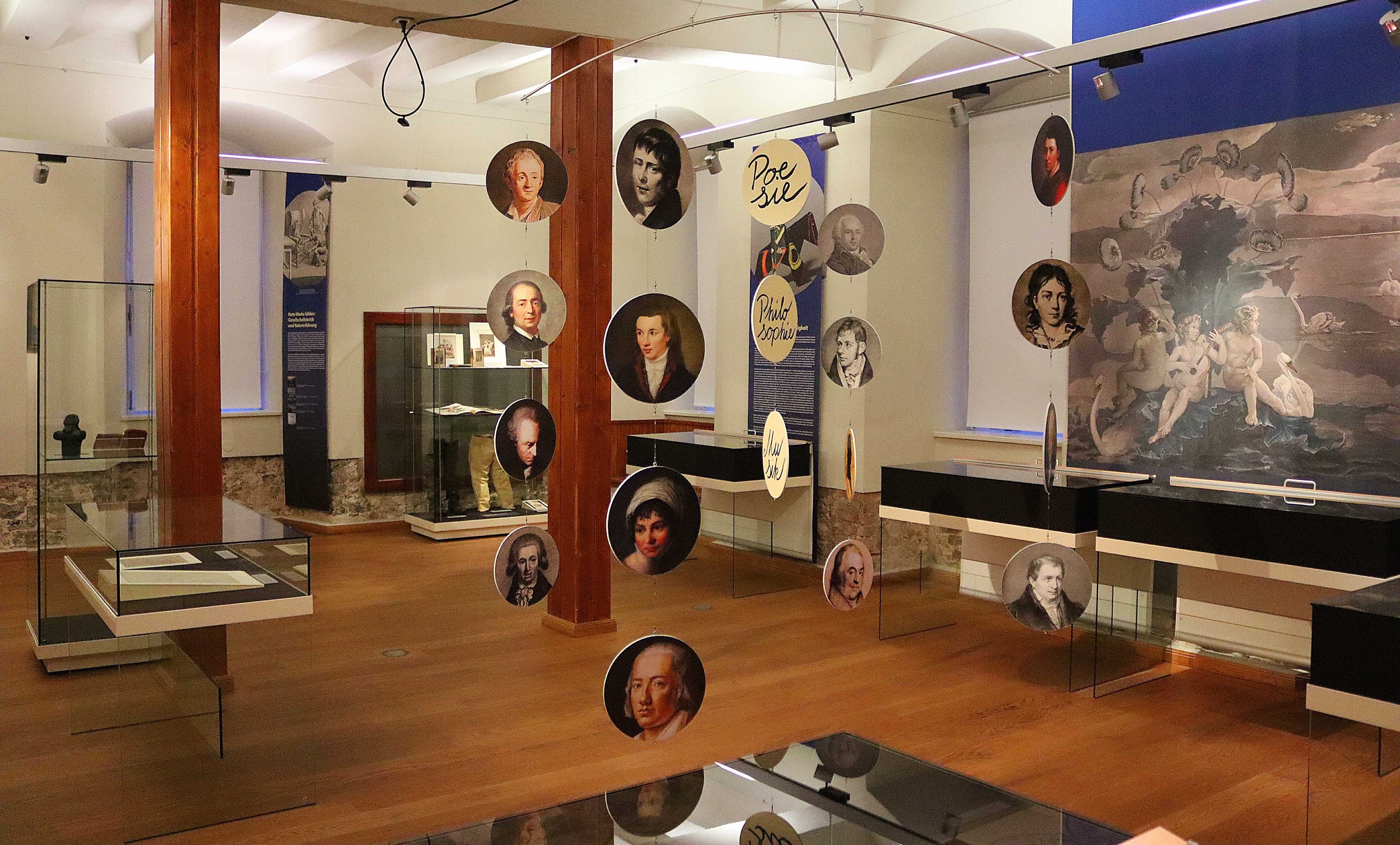 Sonderausstellung "Wie verkörperte Worte". Im Vordergrund hängen runde Porträts untereinander an einem Mobilé. Im Hintergrund stehen verschiedengroße Vitrinen mit Ausstellungsgegenständen.