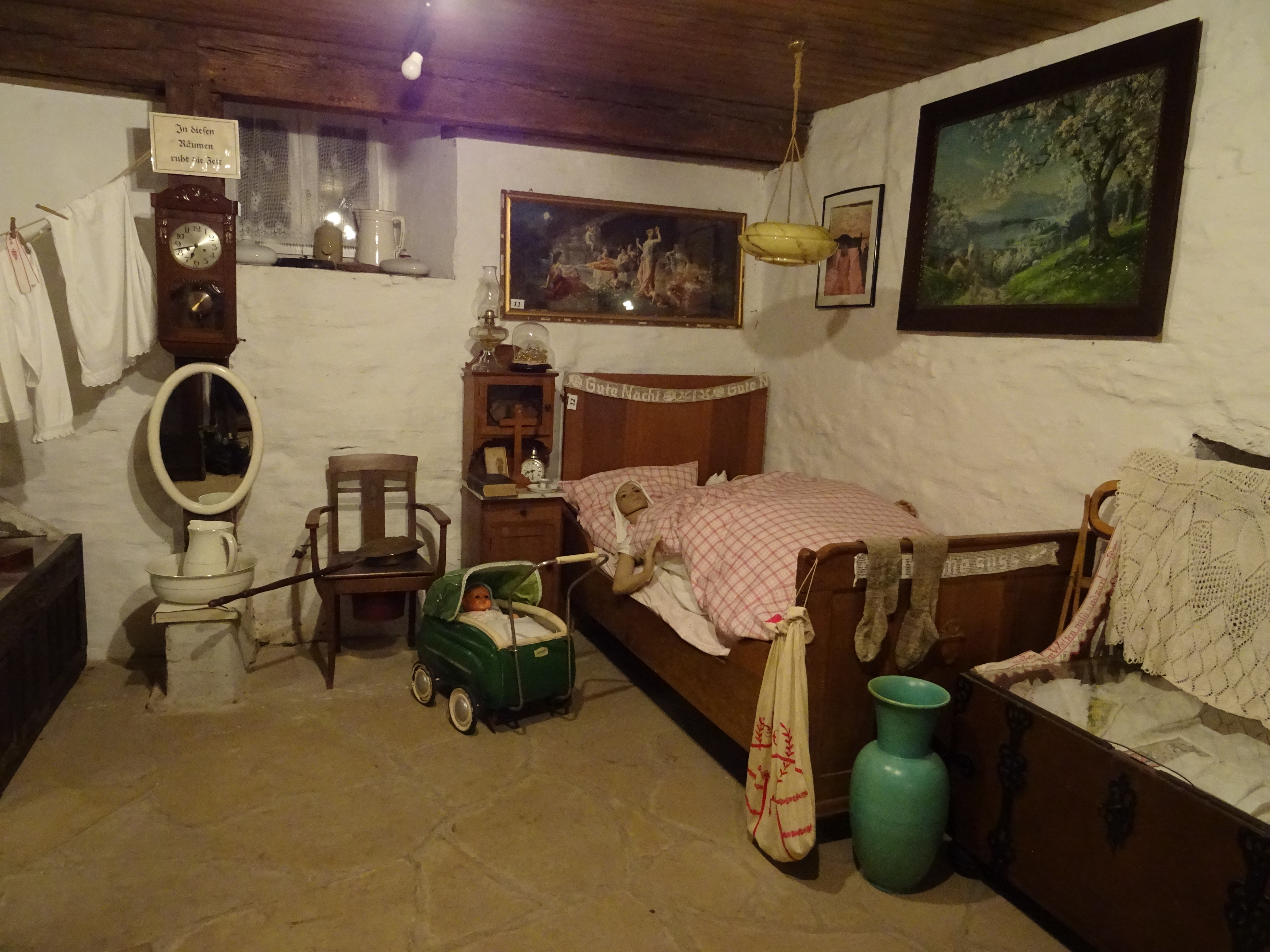 Dieser Raum präsentiert eine Schlafkammer. Bett, Spiegel, aufgehängte Wäsche sowie eine große Truhe, eine Standuhr und Bilder an den Wänden füllen den Raum.