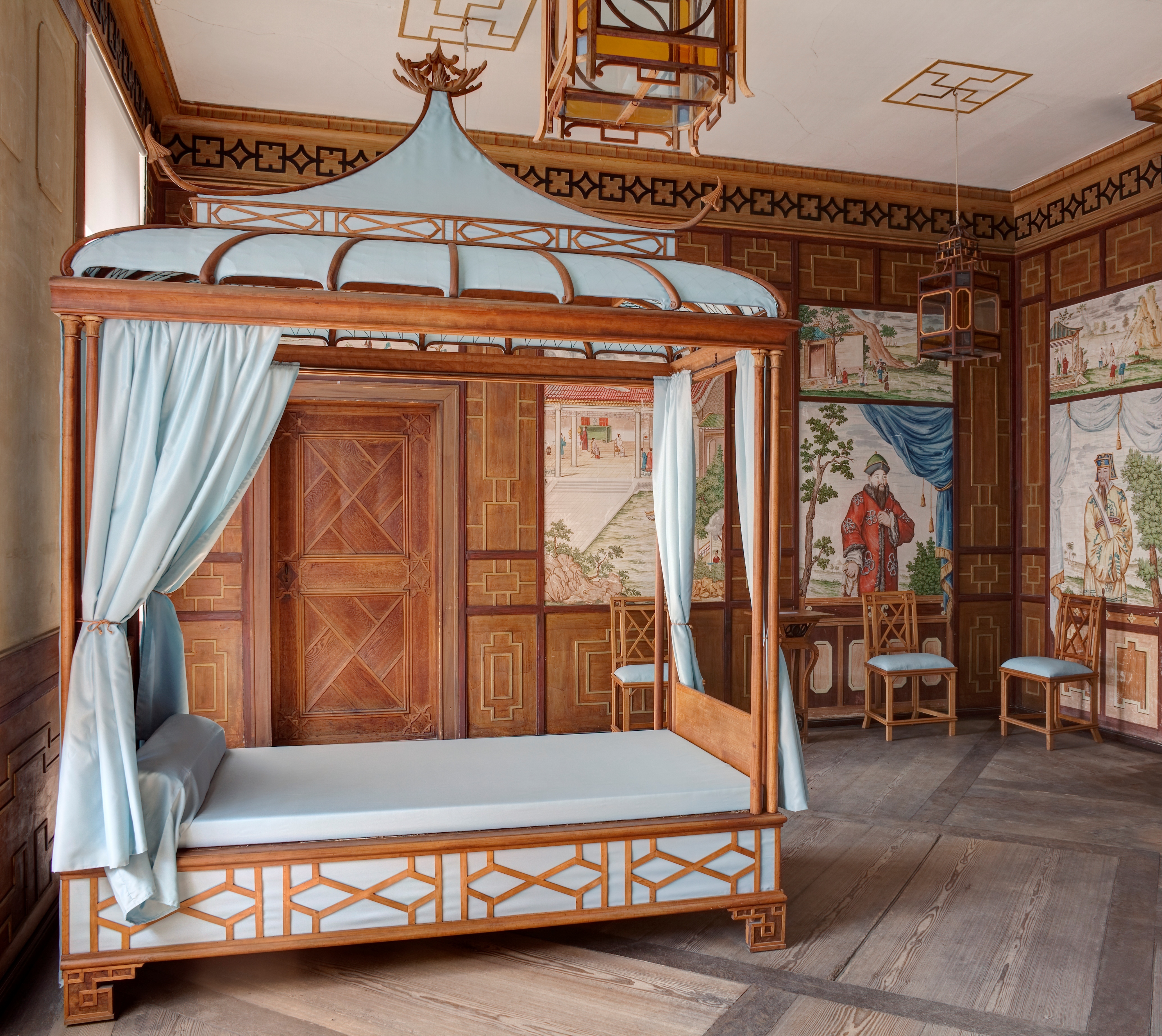 Chinesisches Zimmer im Schloss Oranienbaum. Im Vordergrund steht ein Holzbett mit einem blauen Baldachin darüber. Die Wände sind mit Holzpanelen ausgestattet und zeigen Gemälde chinesischer Gelehrter.